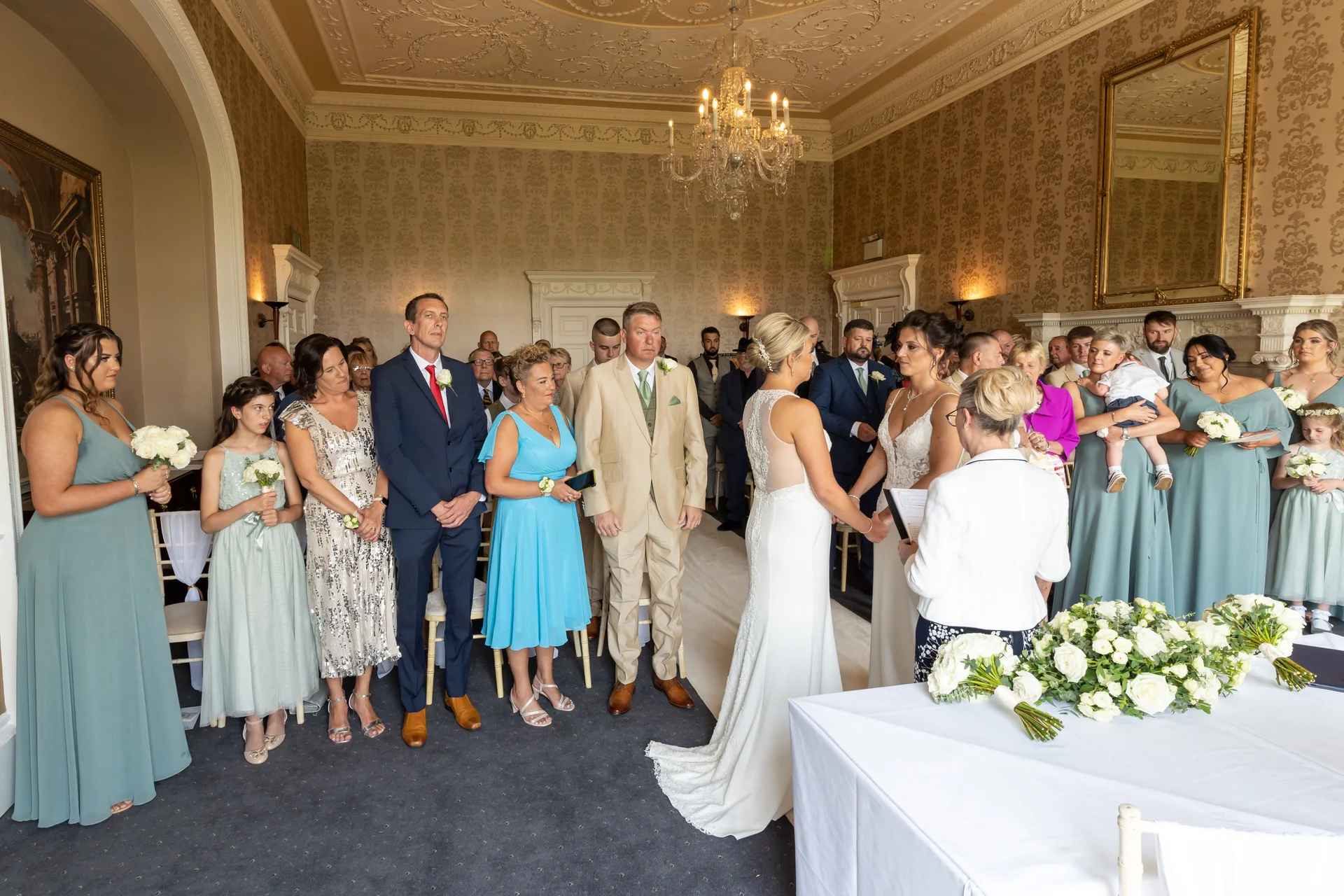 An intimate civil wedding ceremony taking place inside an elegant country house room featuring ornate wall panels and a large chandelier, with the couple exchanging vows while guests look on.