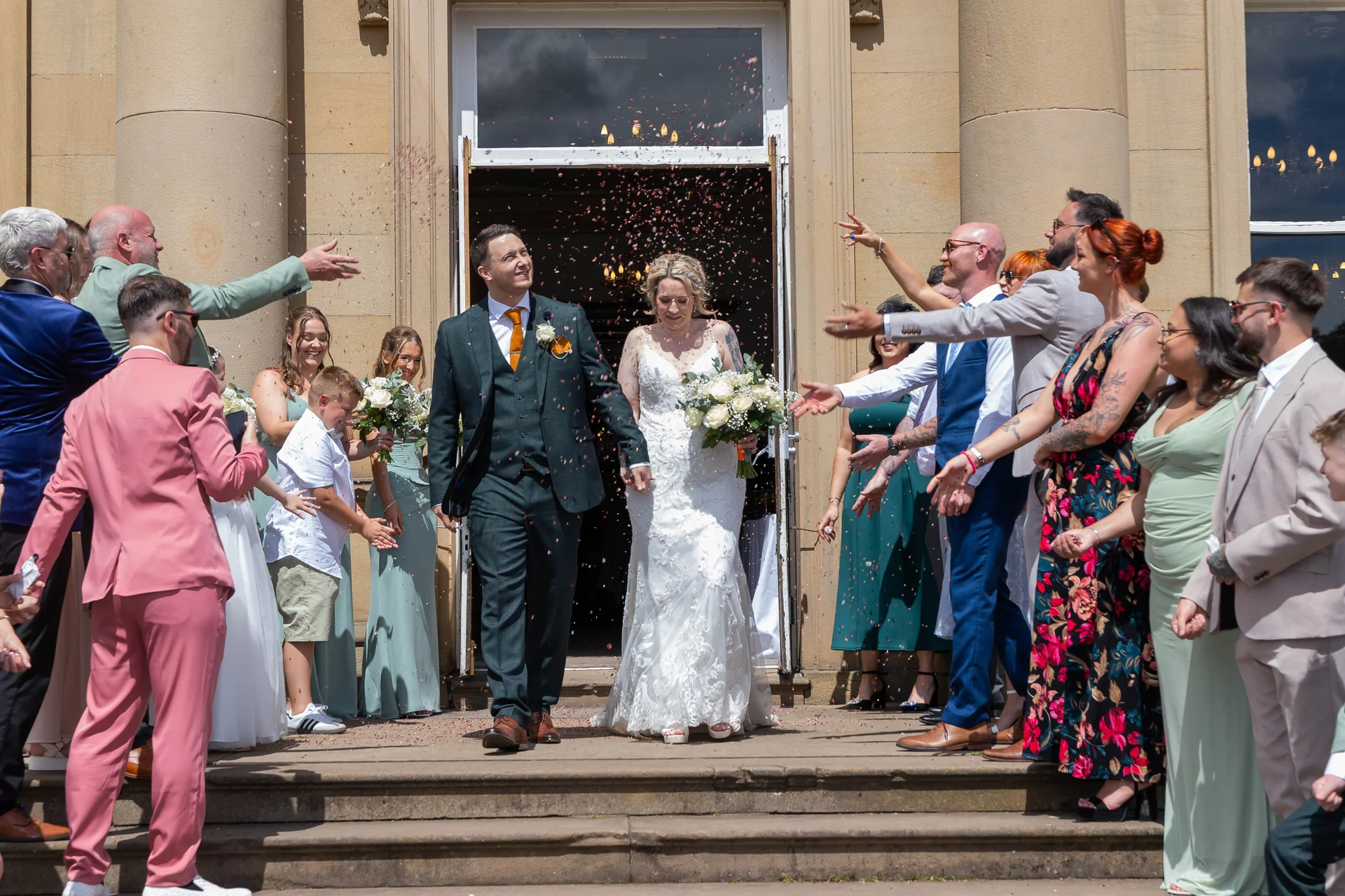 The smiling bride and groom step out of a grand stone doorway as wedding guests eagerly shower them with a cascade of colourful confetti.