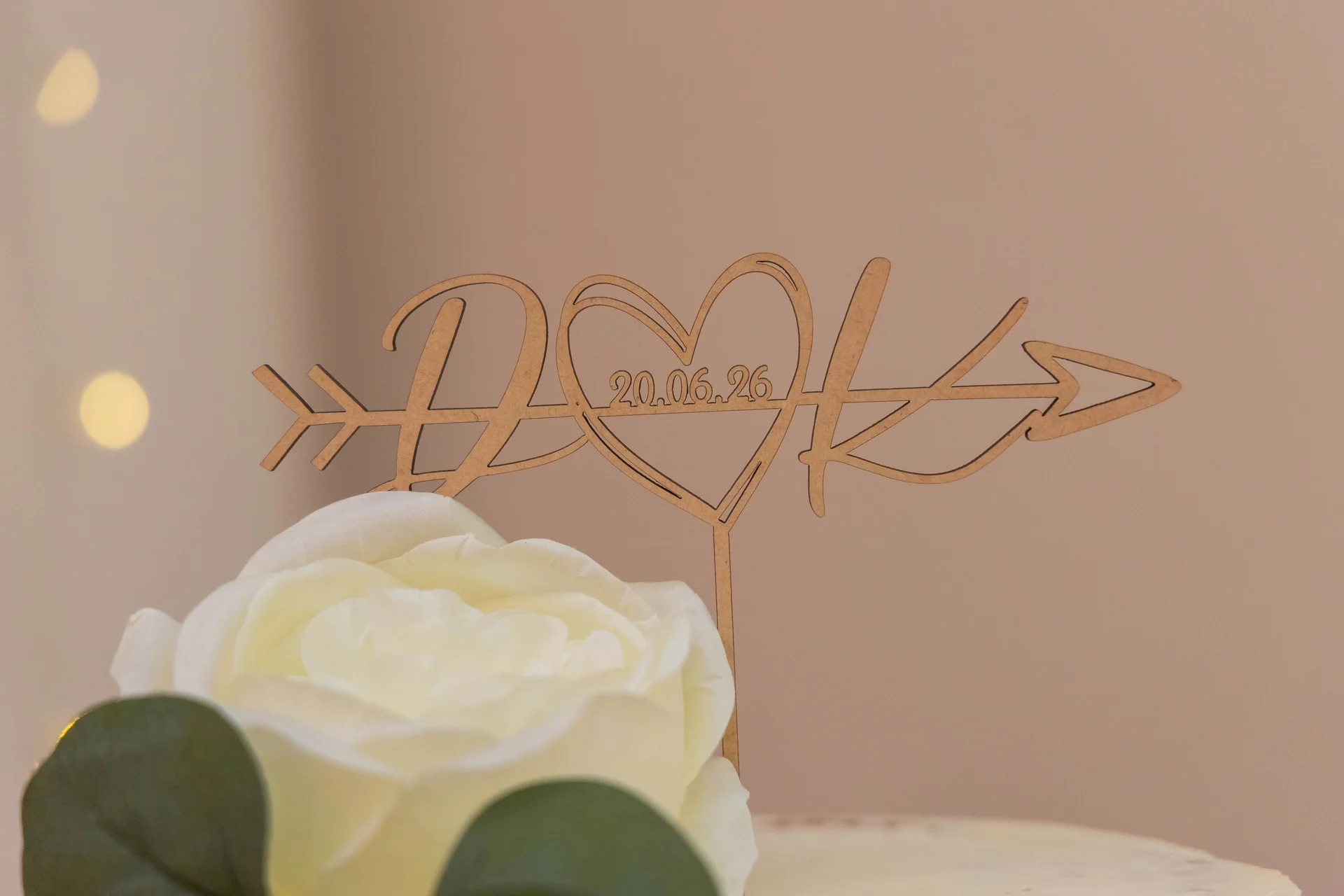 A close-up shot of a custom wooden laser-cut cake topper featuring initials and a wedding date, positioned behind a white rose.