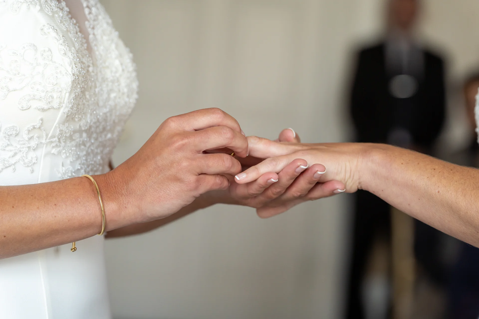 A close-up shot of a bride wearing a beaded white gown gently placing a wedding ring onto her partner's finger during a ceremony.