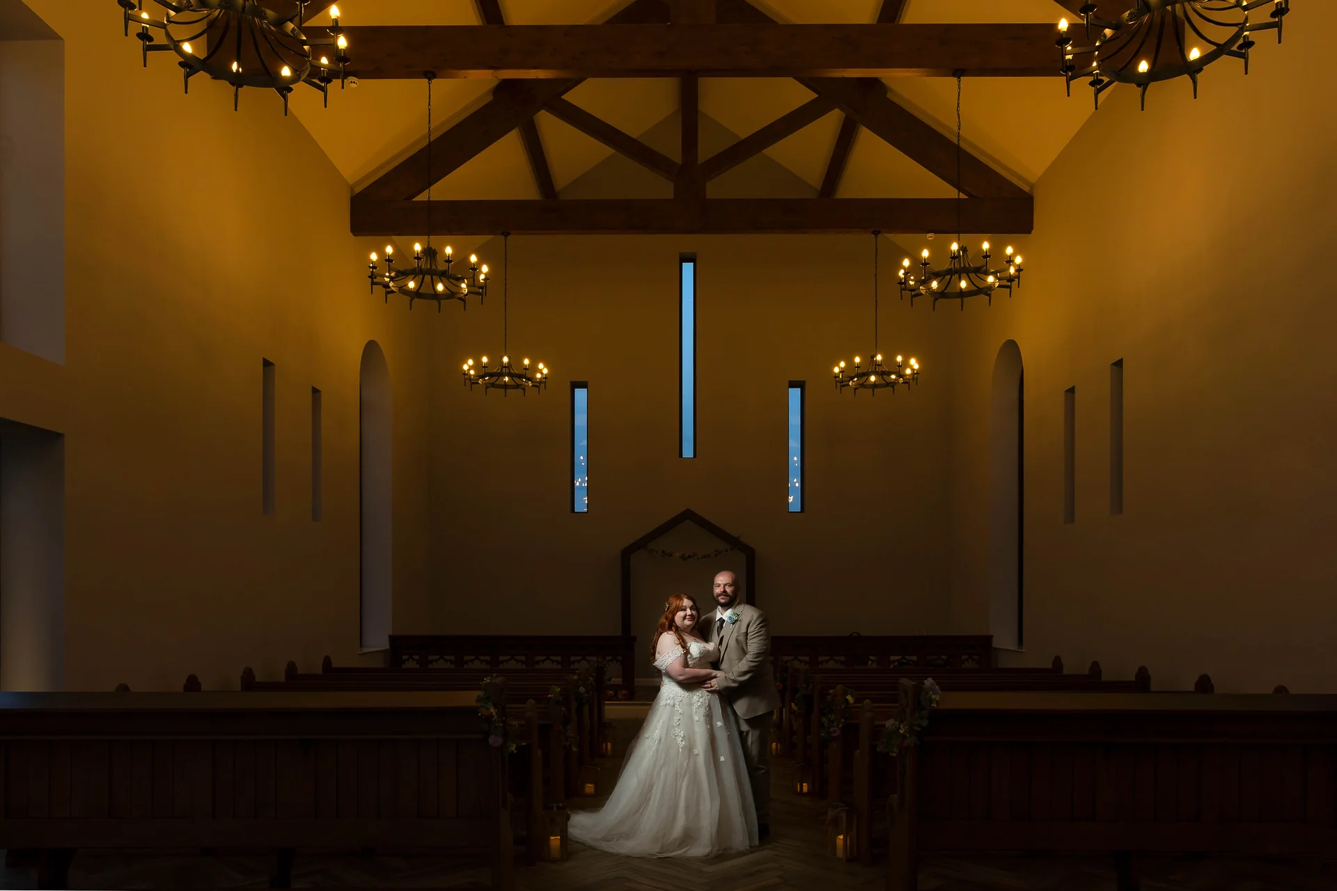 A newlywed couple stands embraced at the aisle of a grand, moody church featuring high vaulted ceilings with exposed timber beams and warm glowing chandeliers.