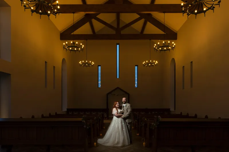 A newlywed couple stands embraced at the aisle of a grand, moody church featuring high vaulted ceilings with exposed timber beams and warm glowing chandeliers.