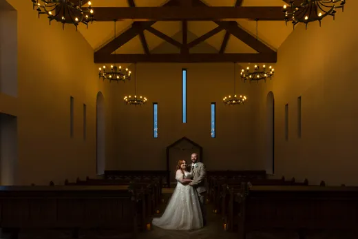 A newlywed couple stands embraced at the aisle of a grand, moody church featuring high vaulted ceilings with exposed timber beams and warm glowing chandeliers.