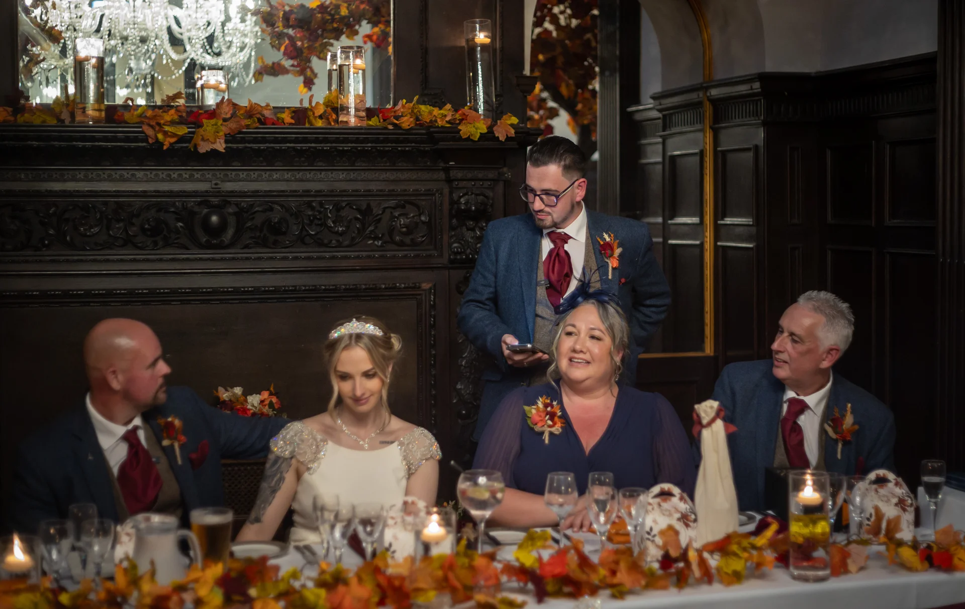 The wedding party gathers at the top table during speeches, surrounded by autumnal leaf decorations and candlelight in a dark wood-panelled room.