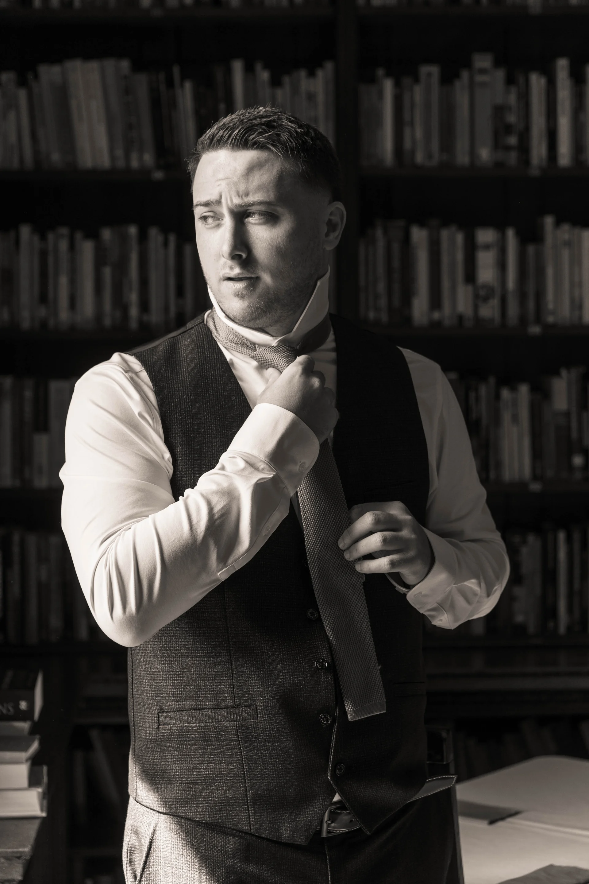 The groom stands in a library adjusting his tie while wearing a waistcoat and shirt.