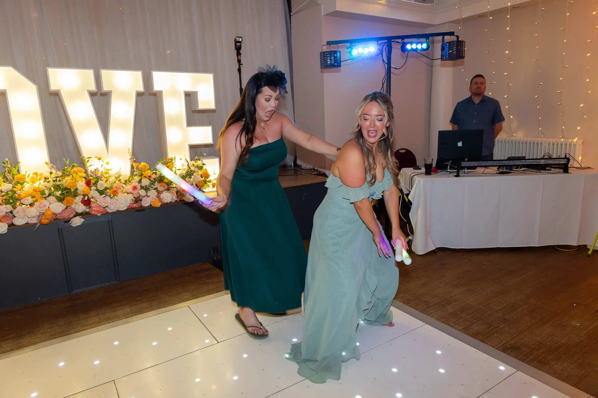 Two bridesmaids enthusiastically dance on an illuminated dance floor holding glowing sticks near large illuminated letters at a wedding reception.