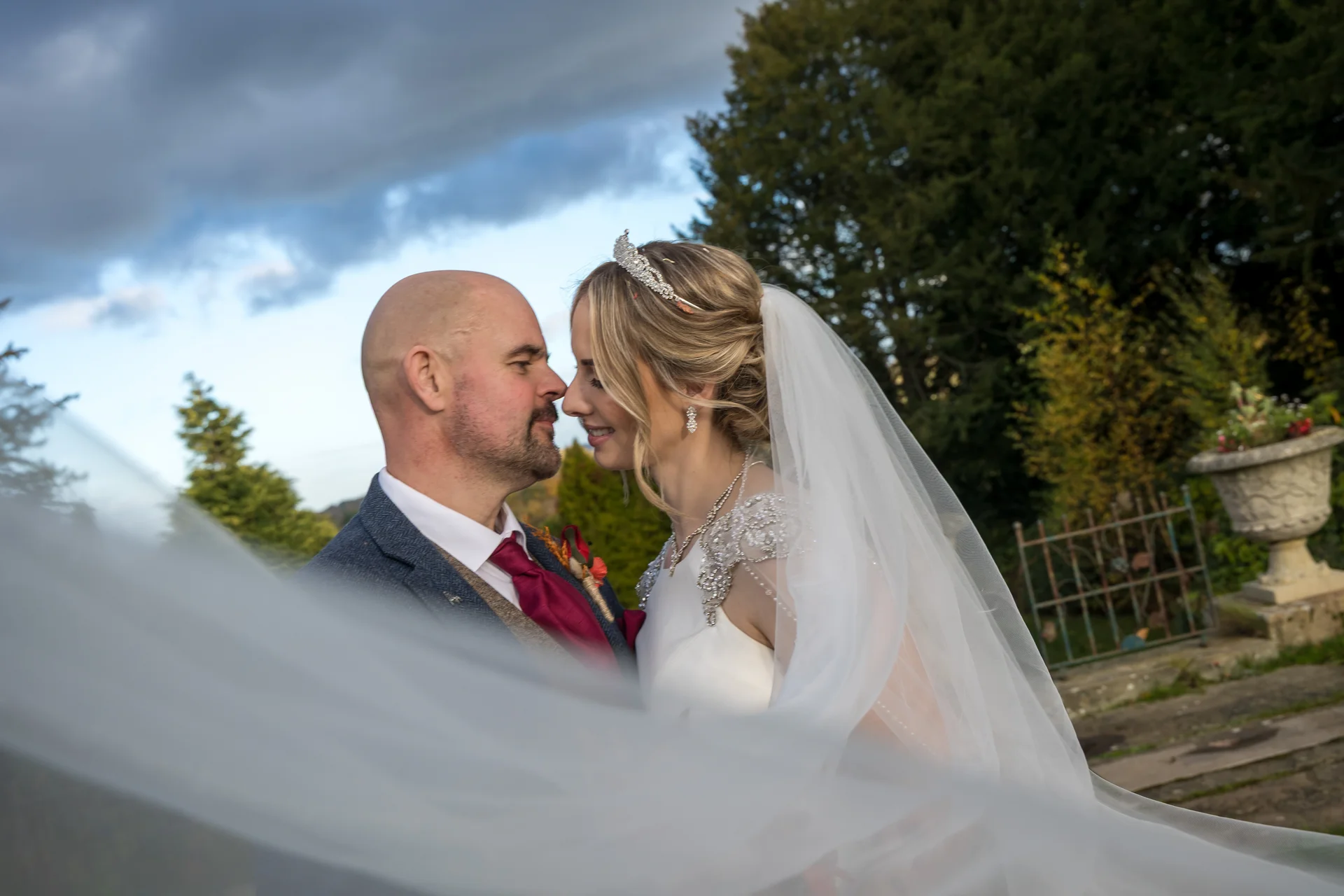 A bride and groom share an intimate moment nose to nose outdoors, framed by a soft tulle veil draped across the foreground against a dramatic evening sky.