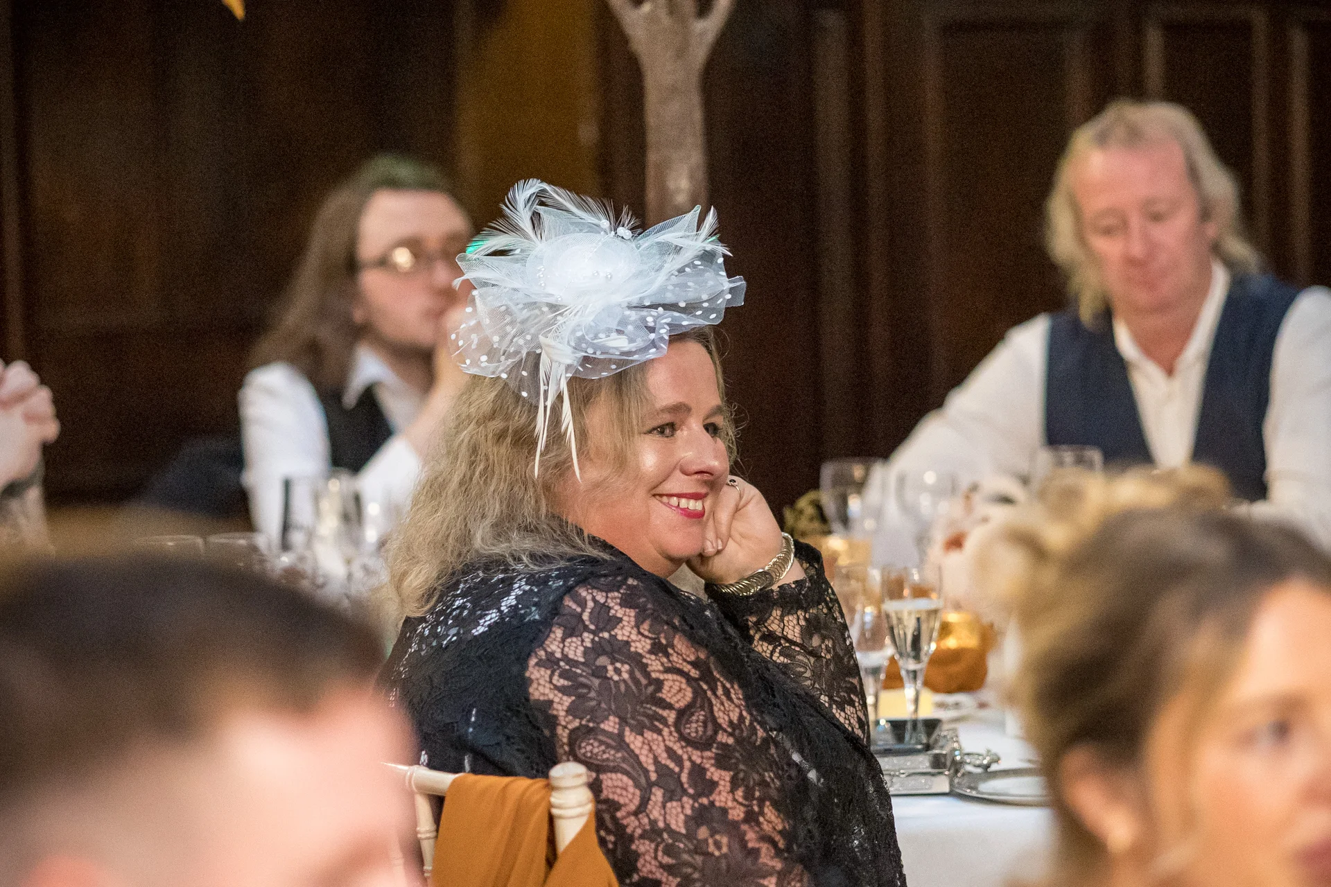 A smiling wedding guest wearing a white fascinator and a black lace dress sits at a banquet table during the wedding reception.