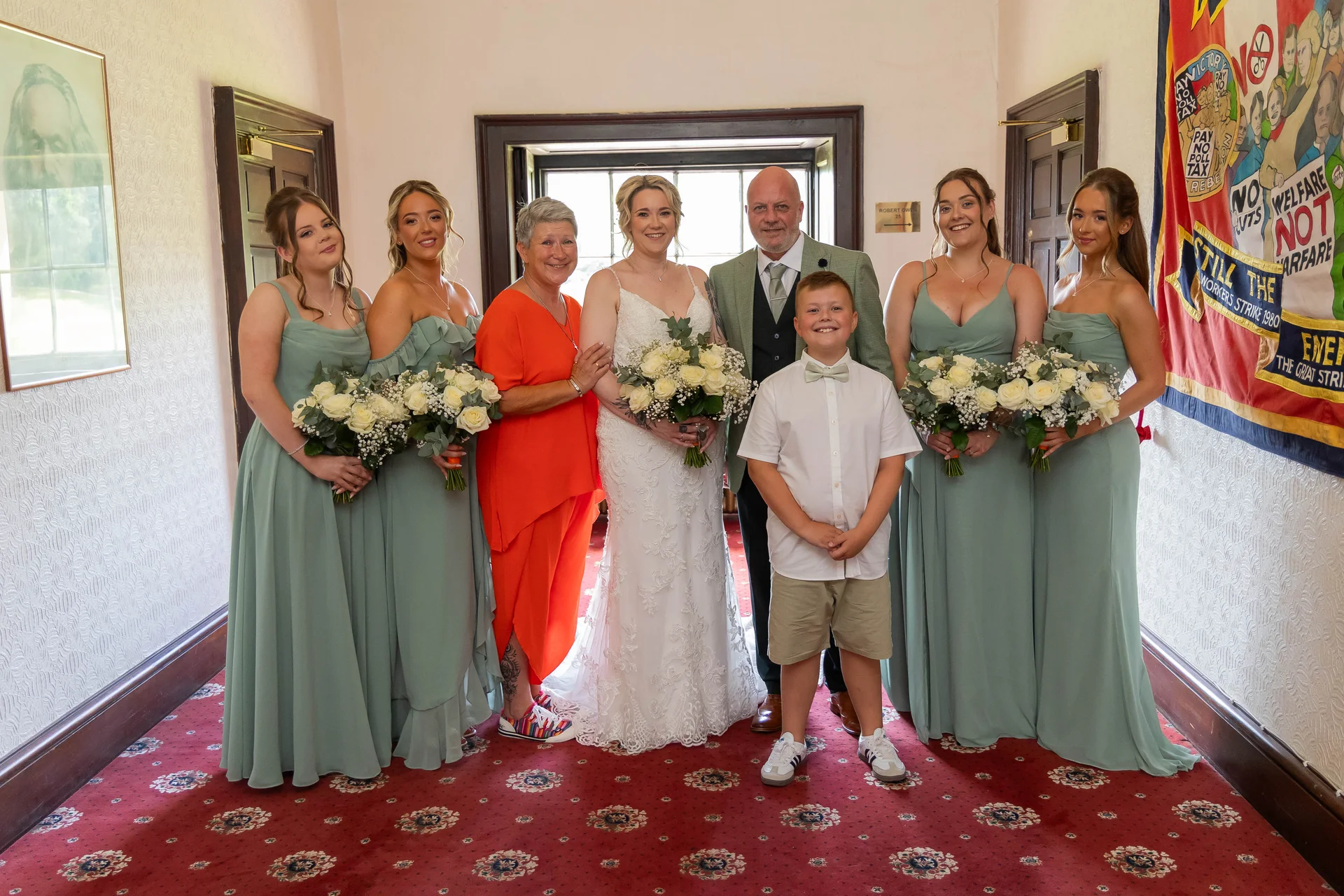 A bridal party poses inside a venue with red patterned carpet, featuring bridesmaids in sage green dresses, the bride in a lace gown, the groom, a young page boy, and a family member in a bright orange outfit.