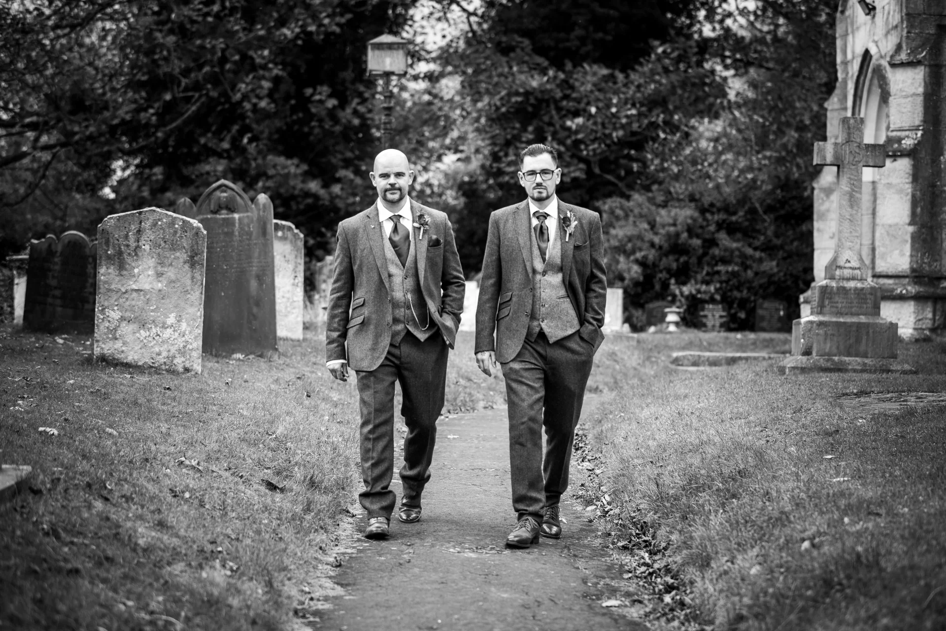 Two grooms in matching tweed suits walk together along a path in a churchyard in a classic black and white photograph.