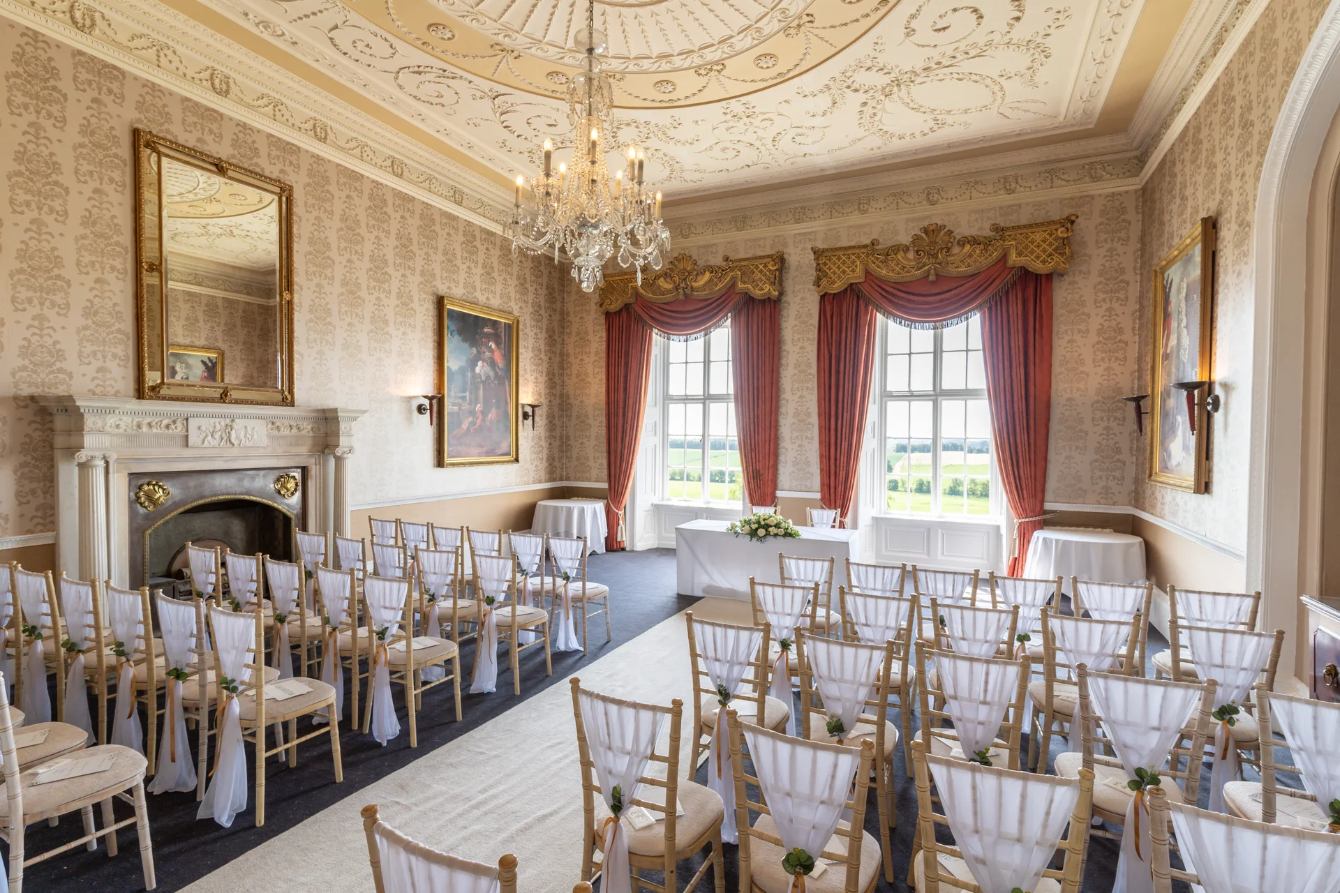 An elegant country house wedding ceremony room features rows of chairs with white sashes facing a clothed table beneath an ornate chandelier and high windows.