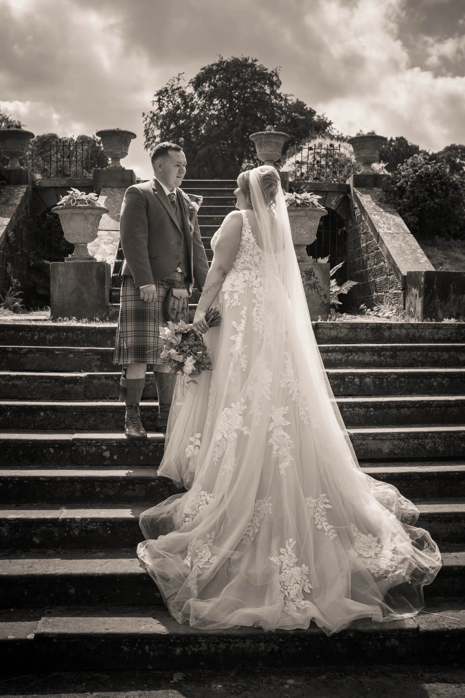 A newlywed couple in sepia tones stand facing each other on grand stone outdoor stairs at a Scottish castle, with the bride wearing an intricate lace gown with a long train and veil.