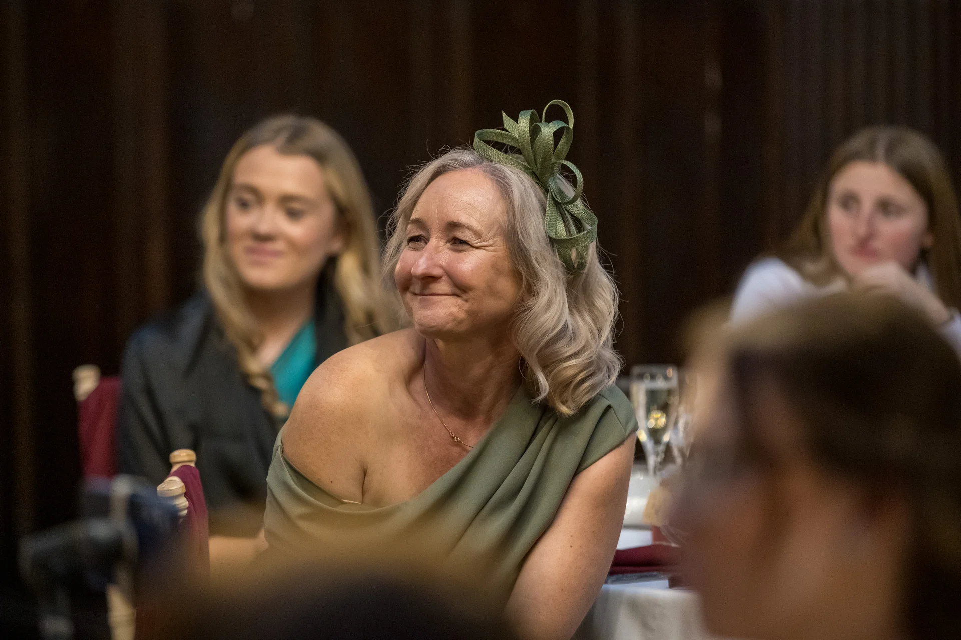 An elegant wedding guest wearing a green dress and matching fascinator smiles warmly while seated at a reception table among other guests.