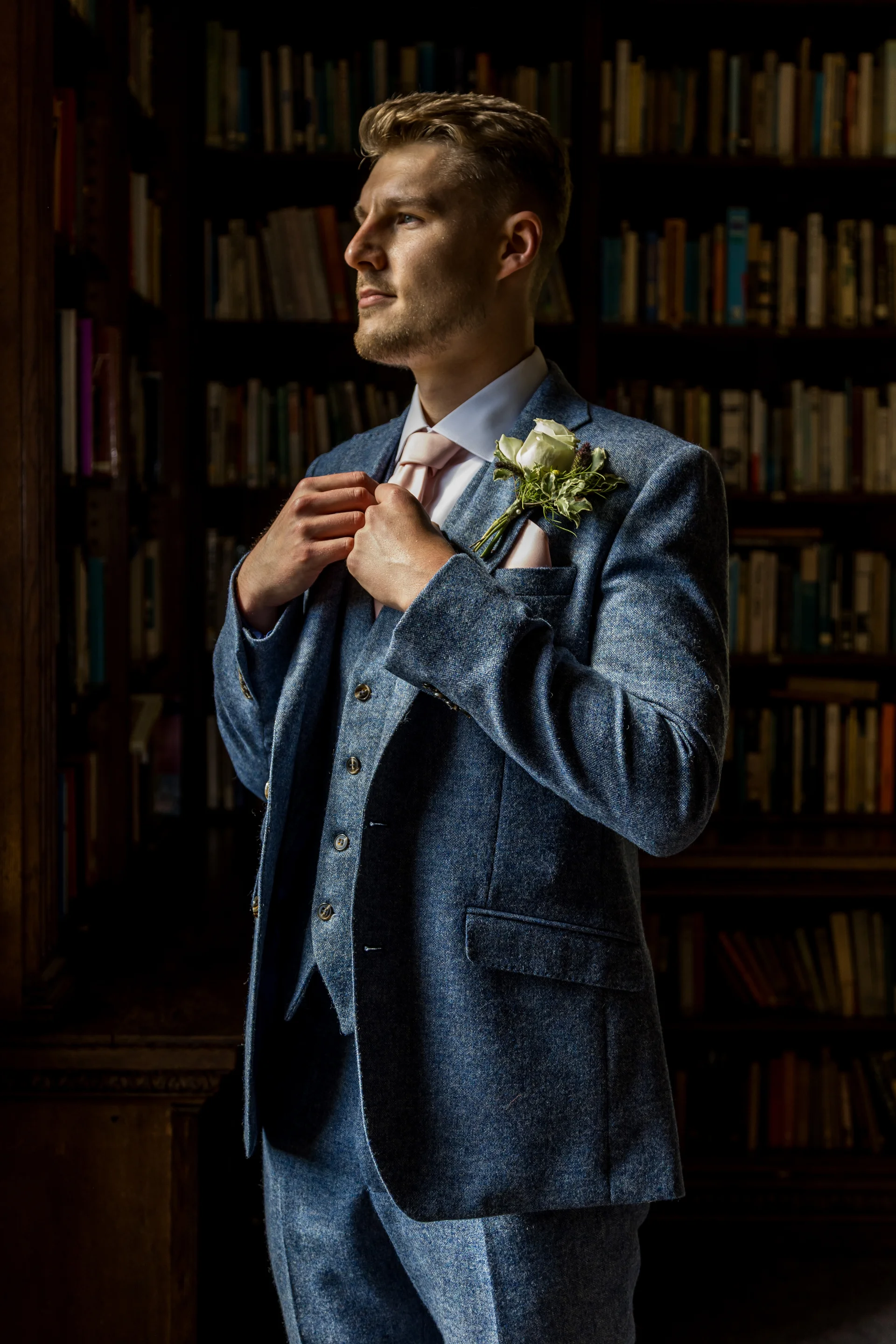 A handsome groom in a blue tweed three-piece suit adjusts his lapel while standing in a dimly lit library filled with floor-to-ceiling bookshelves.