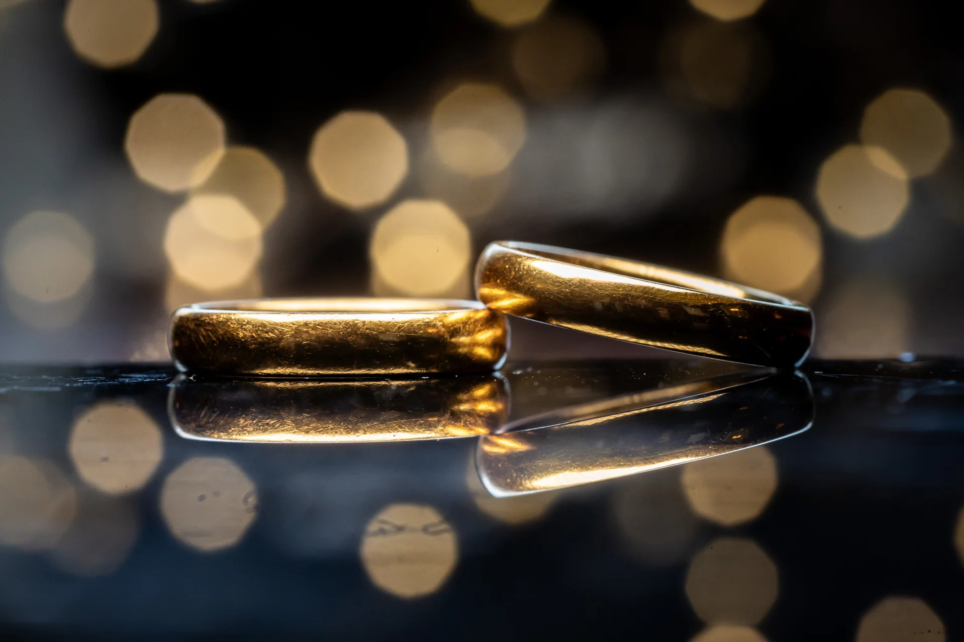 Two gold wedding rings rest on a reflective surface against a backdrop of warm, out-of-focus lights.