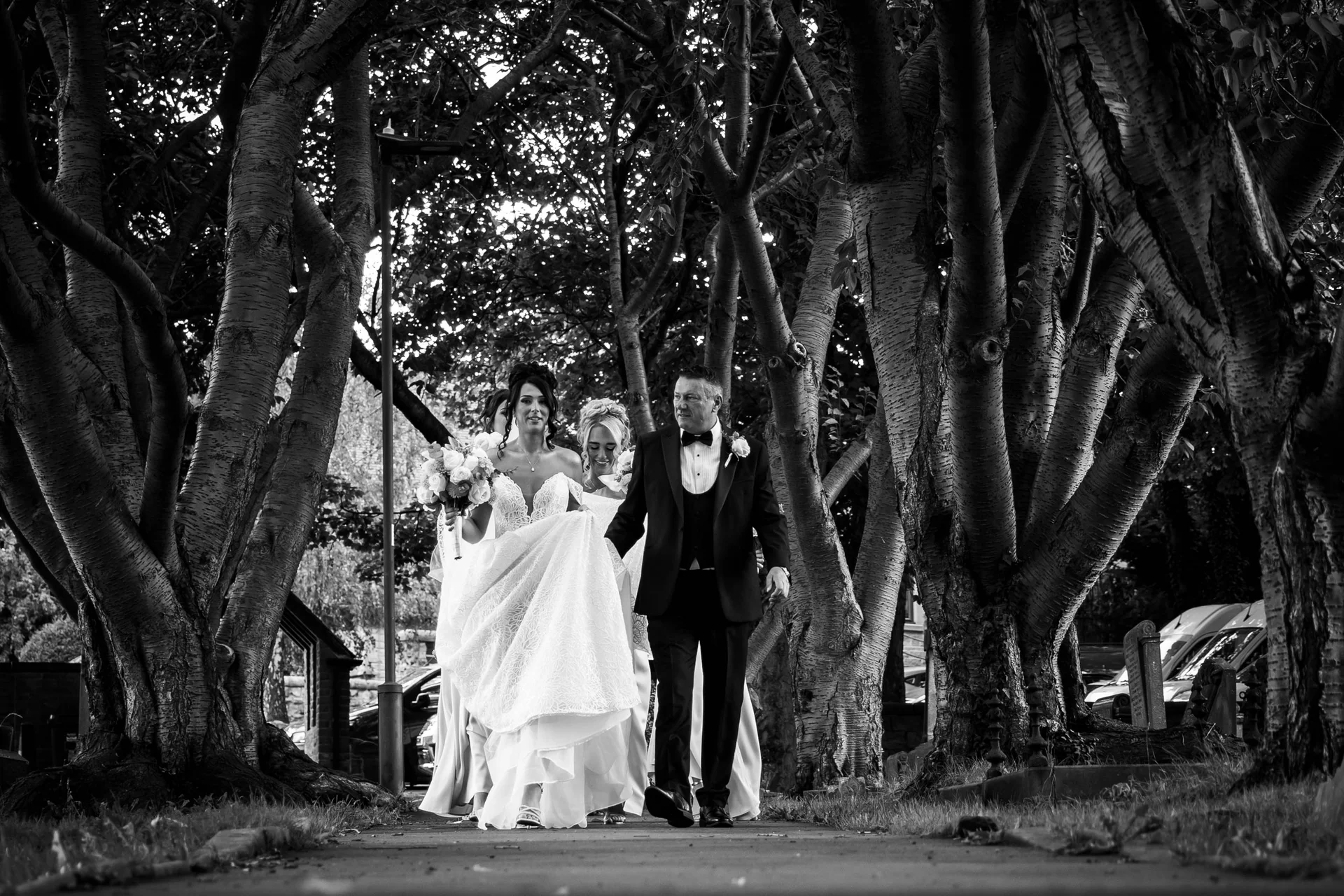 A bride holding her bouquet and a groom in a tuxedo walk together along a path framed by mature trees.