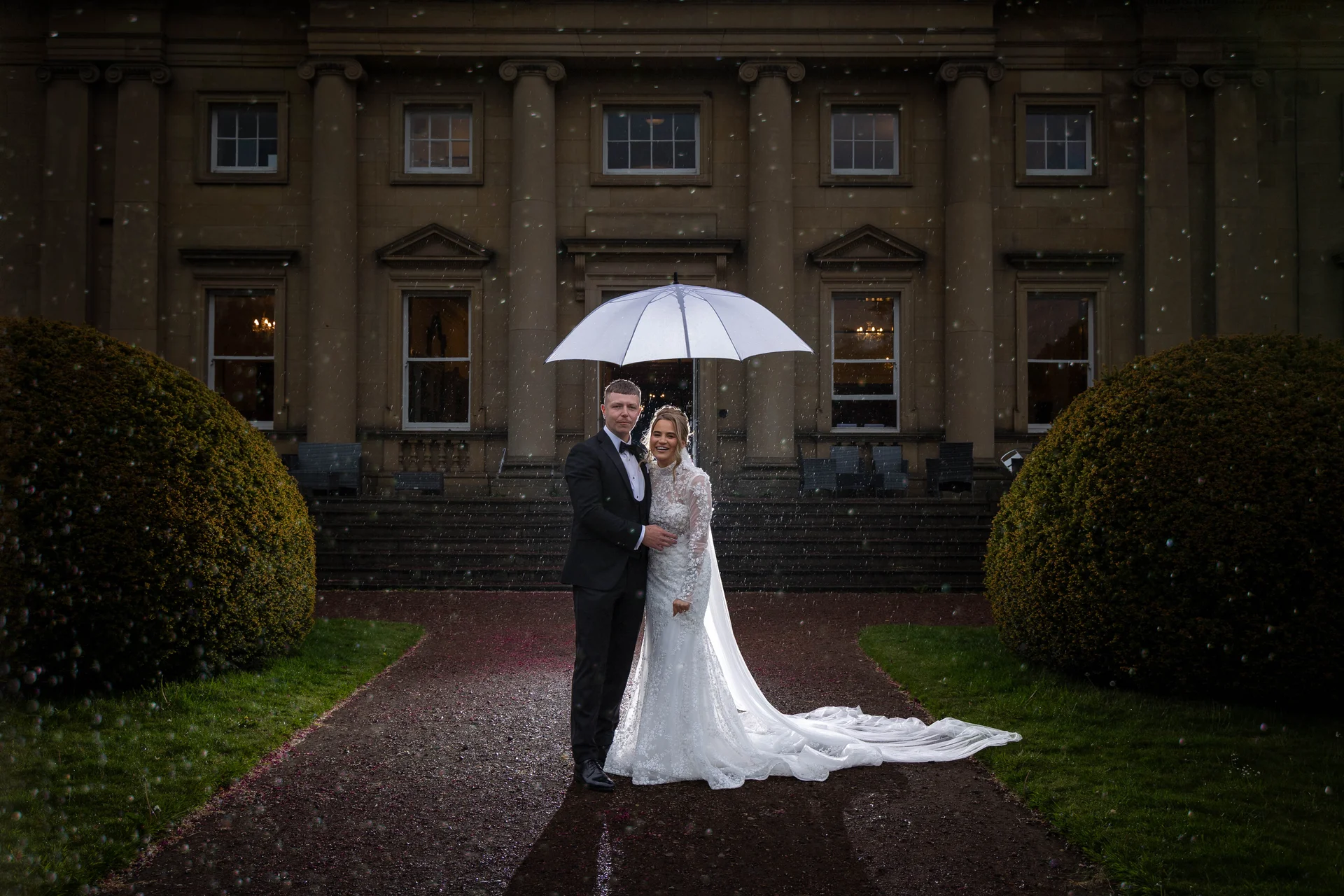 A bride and groom pose happily under a clear umbrella on a wet evening outside a grand country house with illuminated windows.