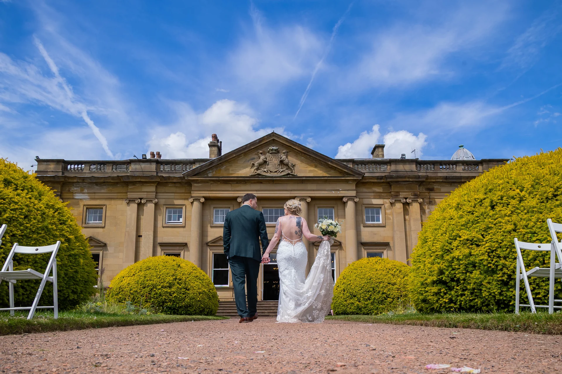 Newlyweds hold hands walking away along a gravel path toward a grand country house under a bright blue sky.