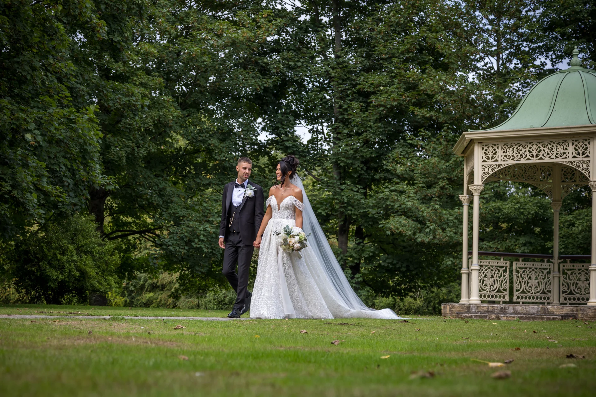 A newlywed couple walks hand in hand across a manicured lawn near an ornate gazebo, the bride wearing an off-the-shoulder lace a-line gown and holding a bouquet while the groom wears a smart black tuxedo.