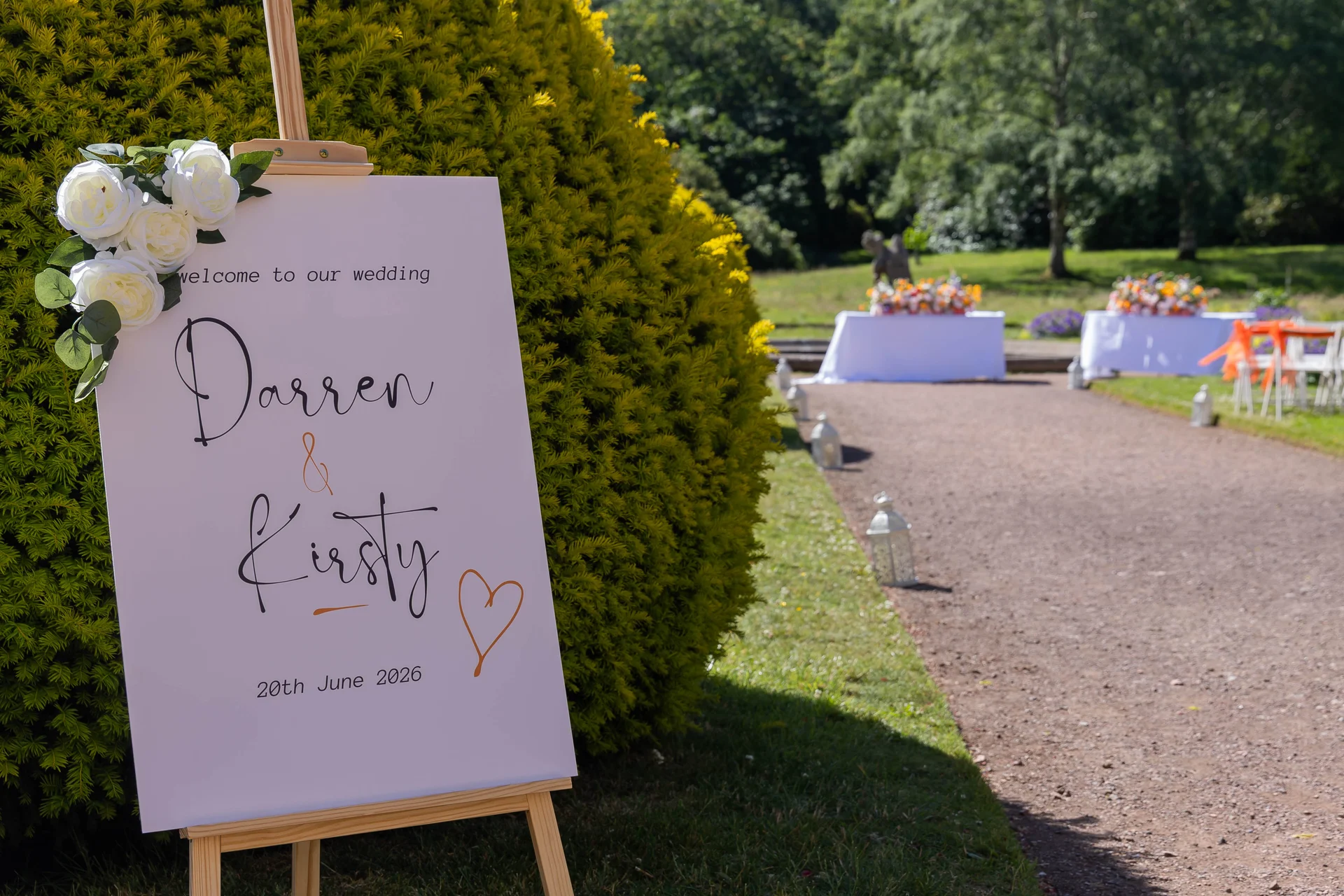 A modern white wedding welcome sign featuring the names Darren and Kirsty stands on a wooden easel beside a path in a manicured garden.