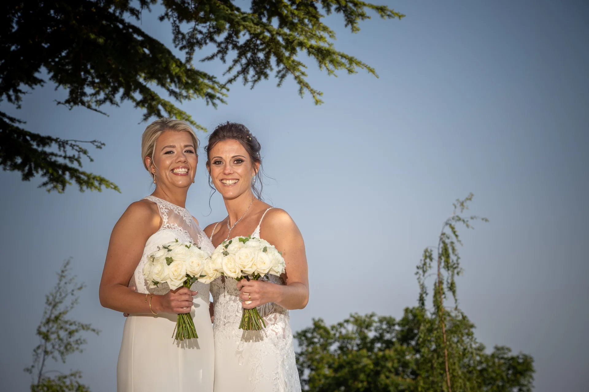 Two smiling brides stand outdoors against a clear blue sky, each holding a hand tied bouquet of white roses.