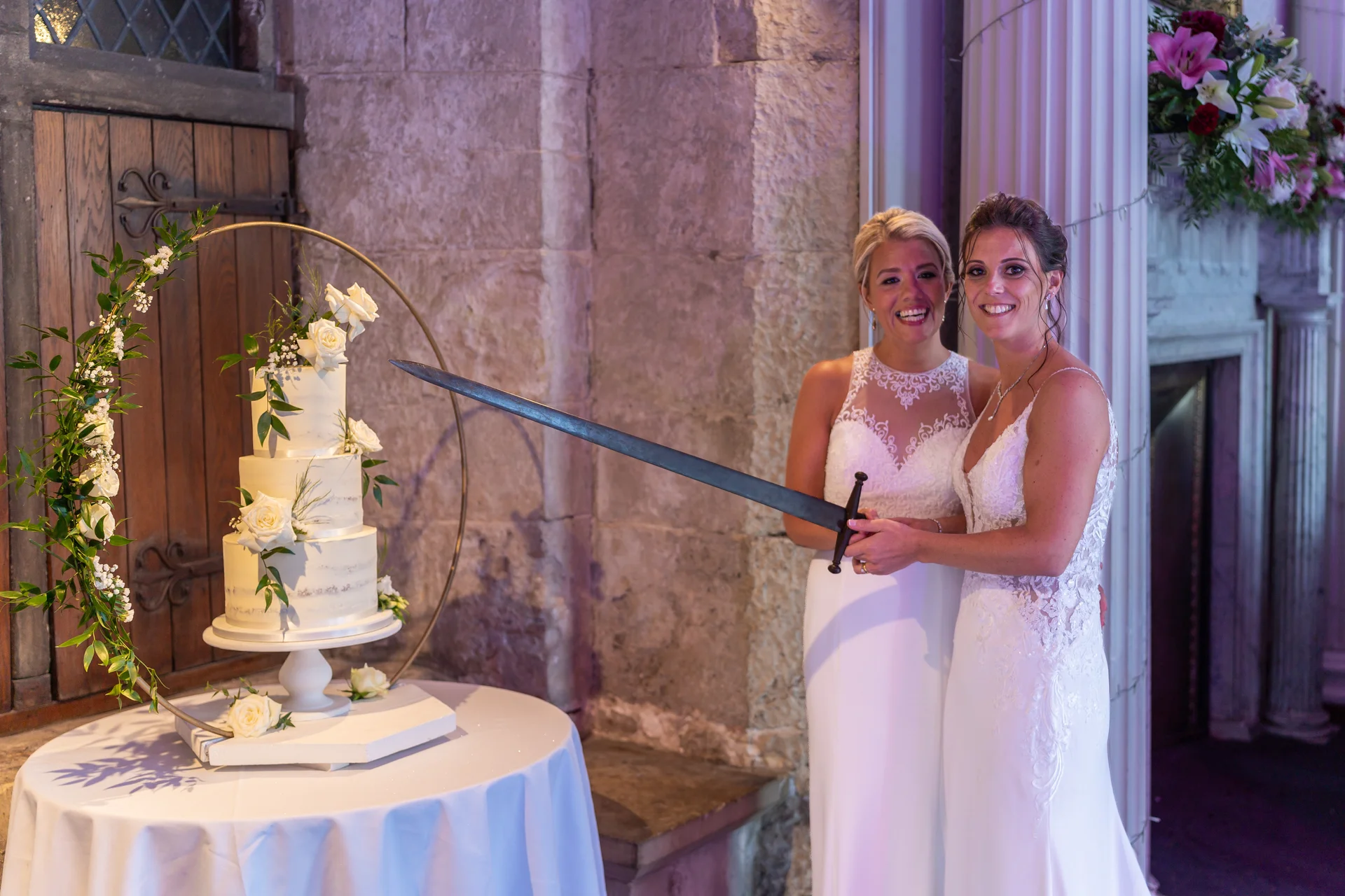 Two smiling brides stand side by side holding a sword together to cut a three-tiered wedding cake adorned with white roses in front of a stone wall.