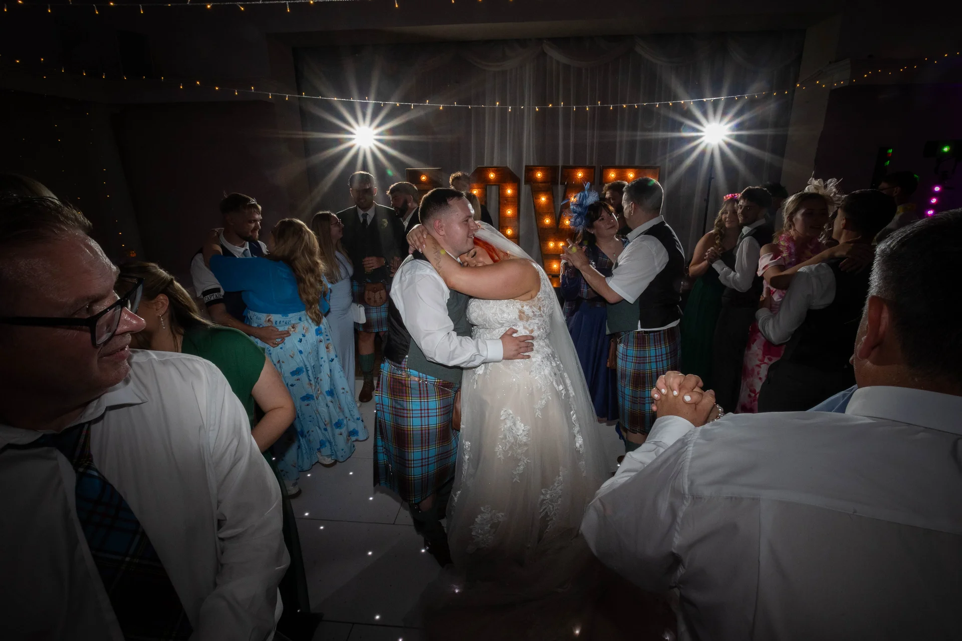 A bride and groom share a tender embrace during their first dance on a lighted dance floor, surrounded by guests in a festive evening reception setting.