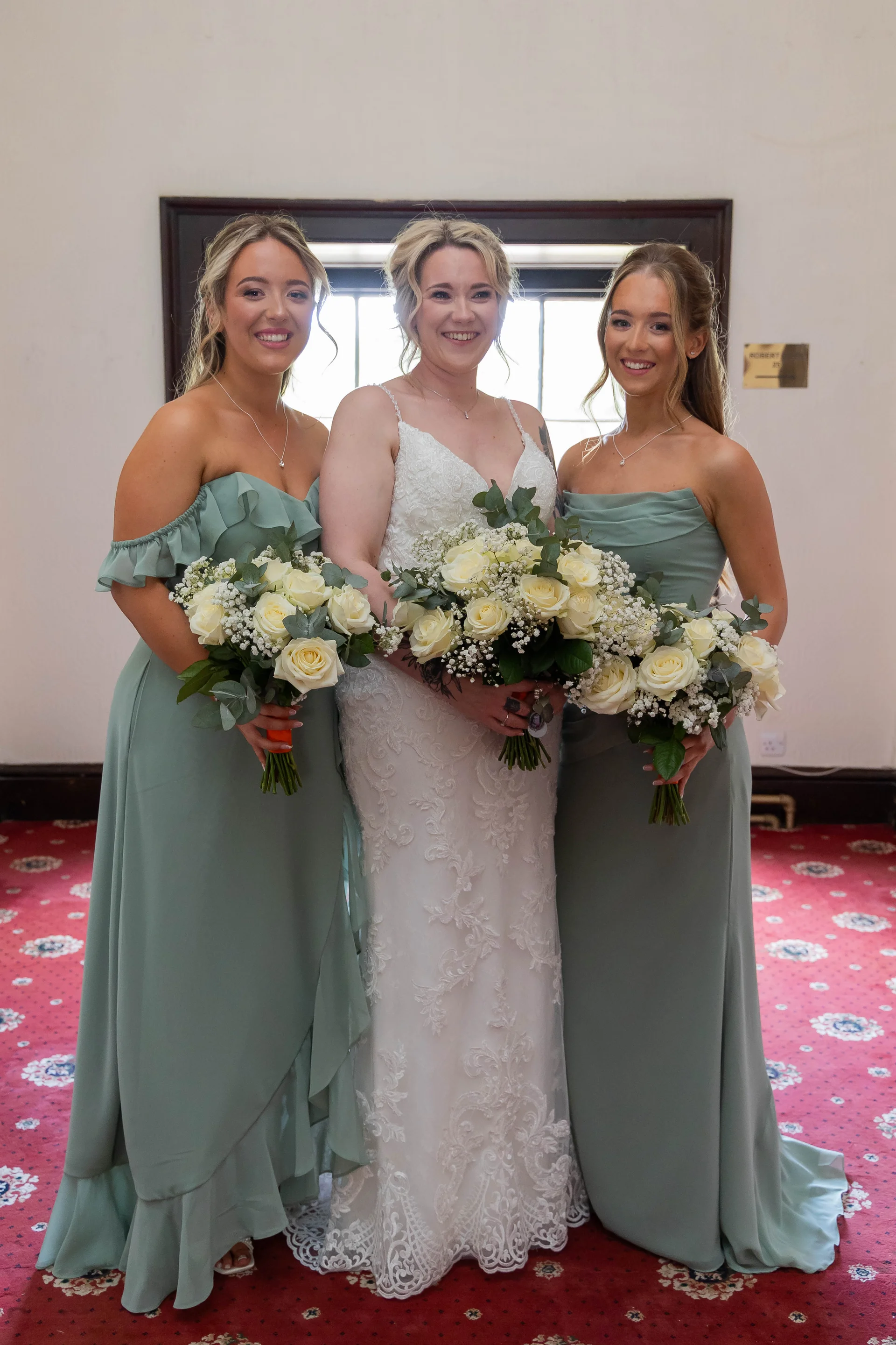 A smiling bride in a white lace a line gown stands between two bridesmaids wearing sage green dresses, each holding a hand-tied bouquet of white roses and greenery.