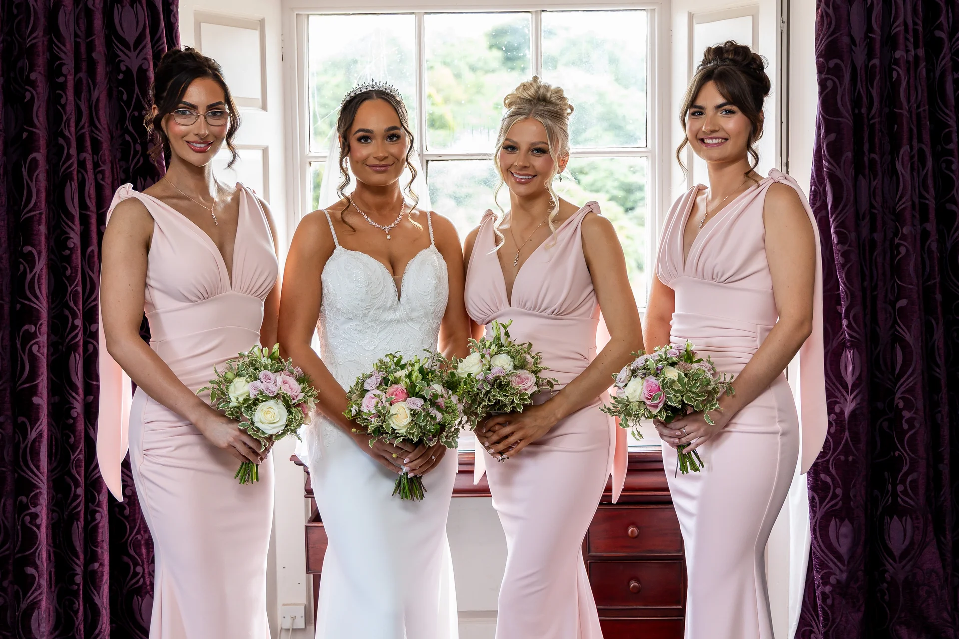 A smiling bride wearing a white gown and tiara poses by a window with her three bridesmaids in matching pink dresses, each holding a floral bouquet.