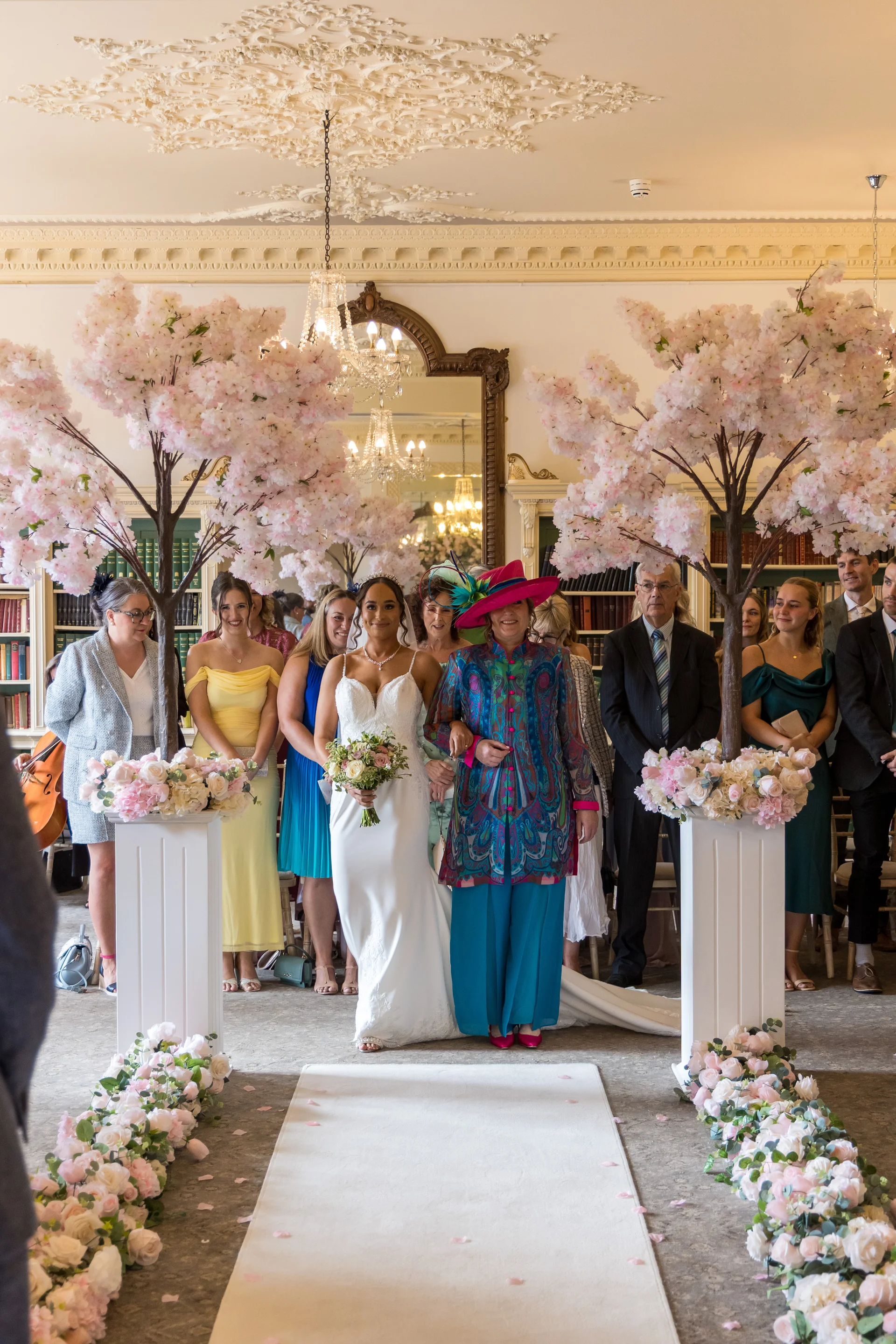 A smiling bride walks down the white aisle runner flanked by cherry blossom trees and floral arrangements, accompanied by her mother in a vibrant blue coat and pink hat.