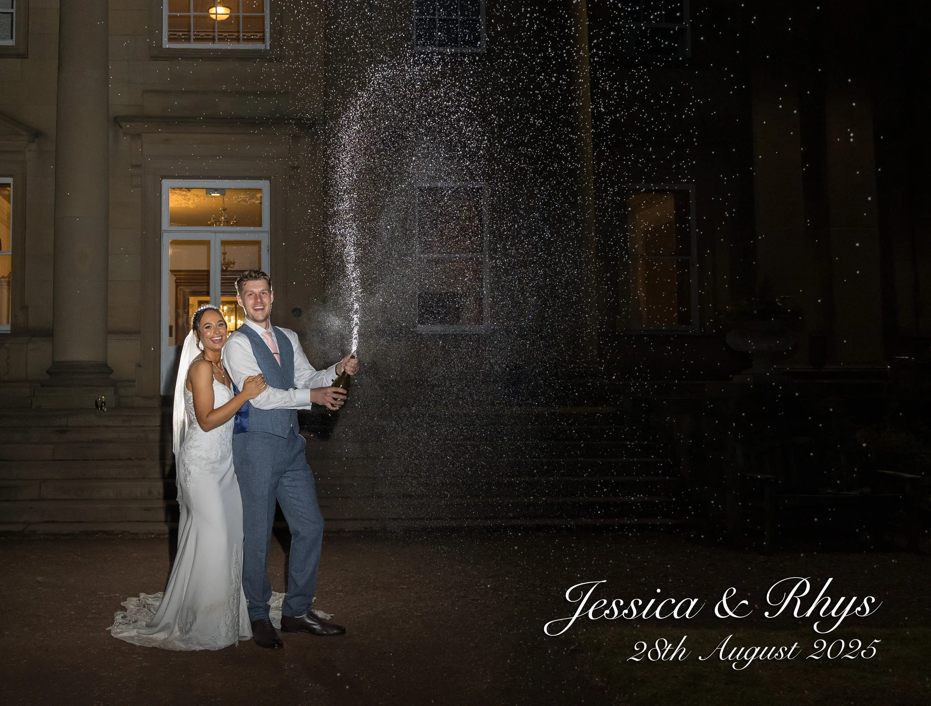 A joyful newlywed couple spraying champagne outside a grand manor house at night, illuminated by dramatic flash photography.