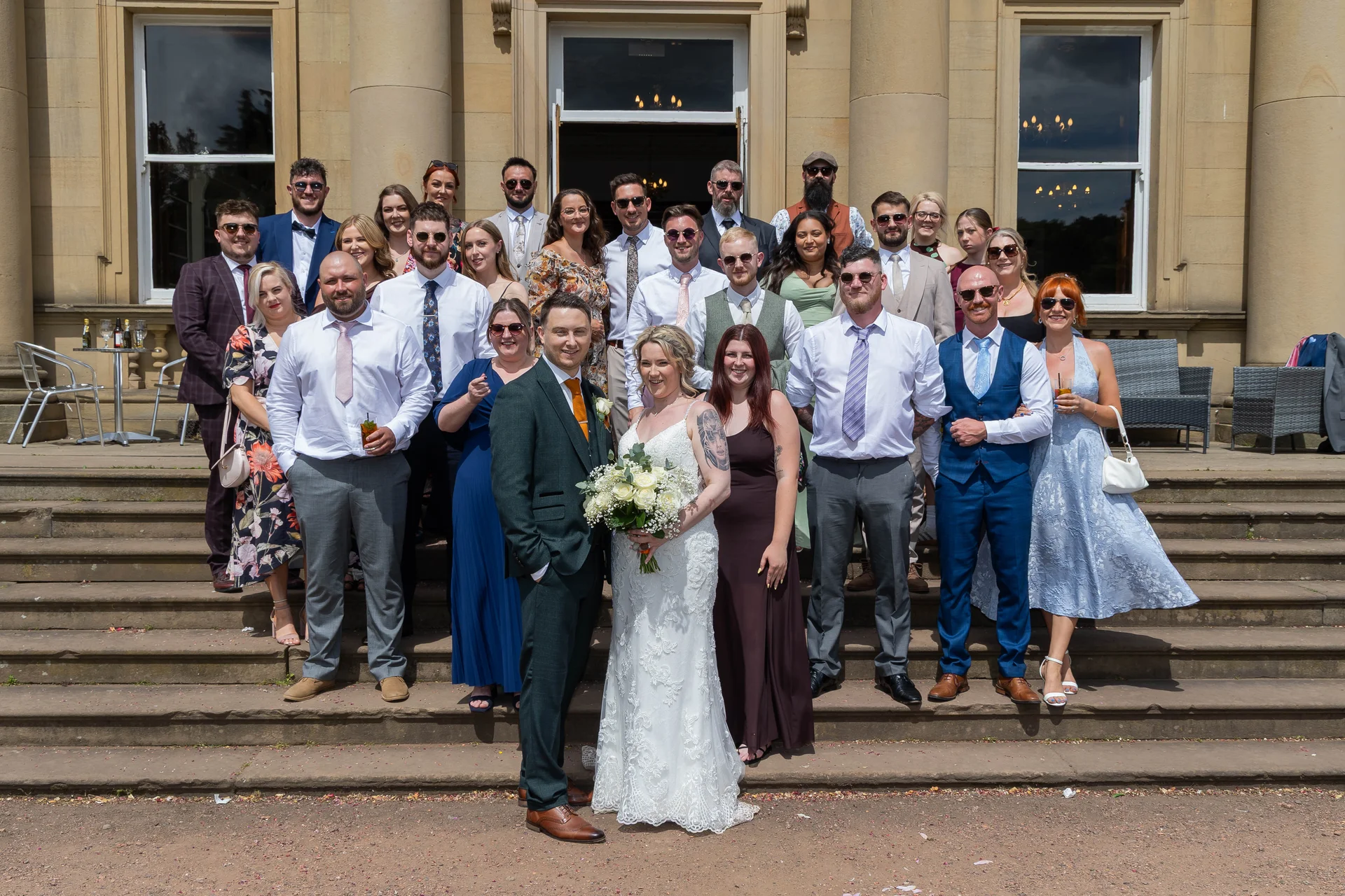 The bride and groom pose with their large wedding party and guests on the grand stone steps outside a stately home under bright sunshine.