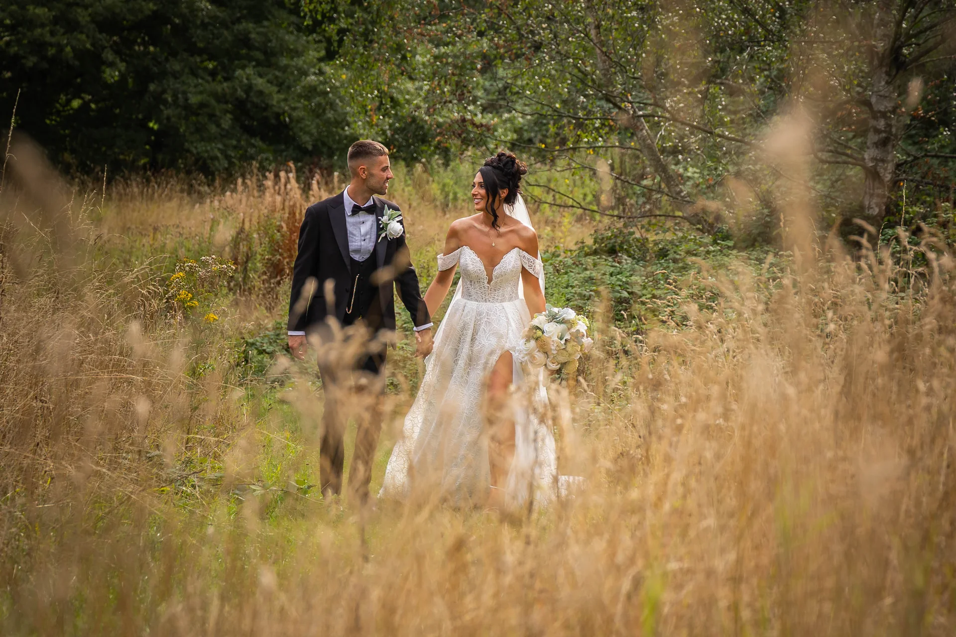 A smiling bride and groom holding hands as they walk through a tall wildflower meadow at the edge of a woodland.