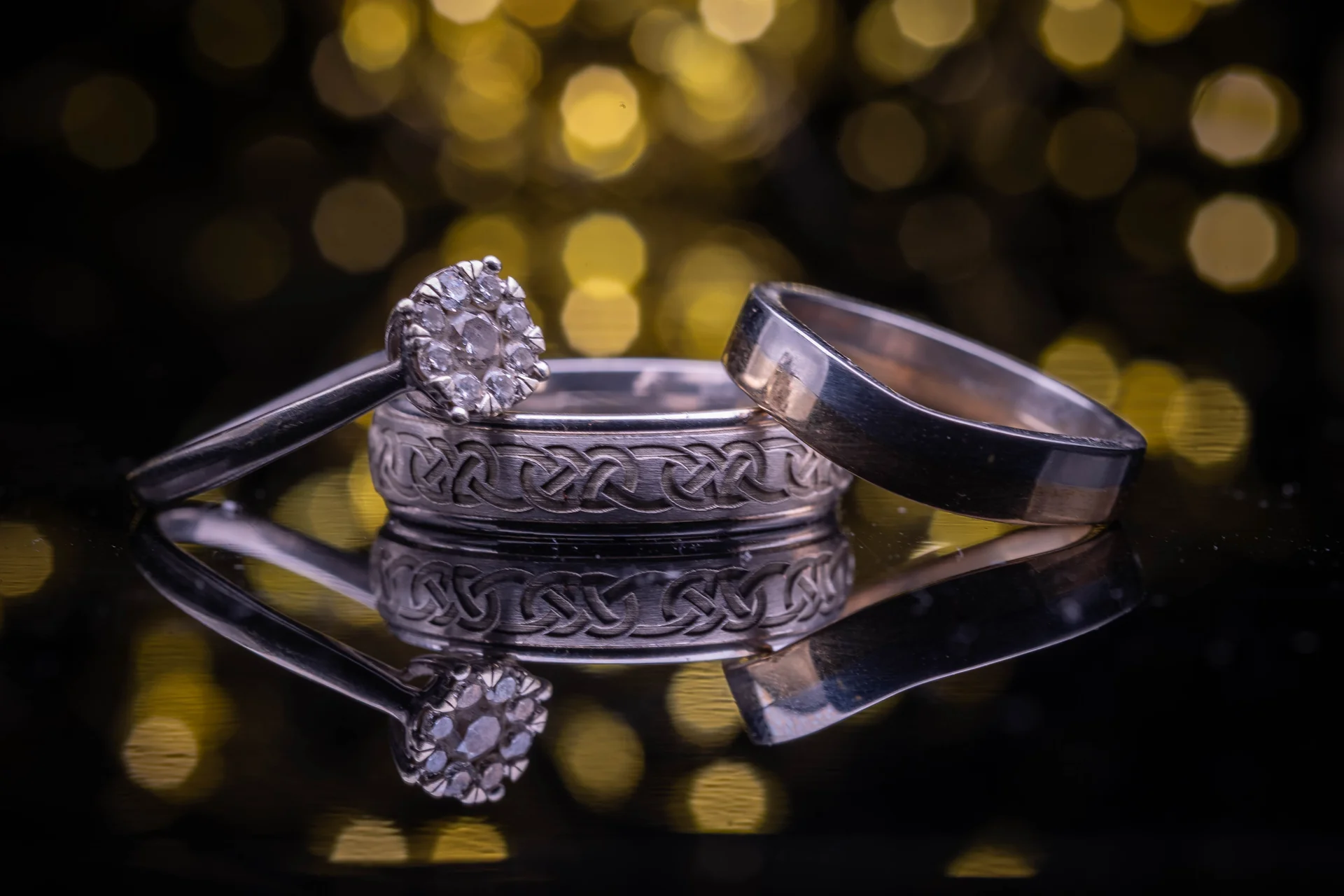Macro detail shot of a diamond engagement ring and matching wedding bands resting on a reflective surface against a glowing golden bokeh background.