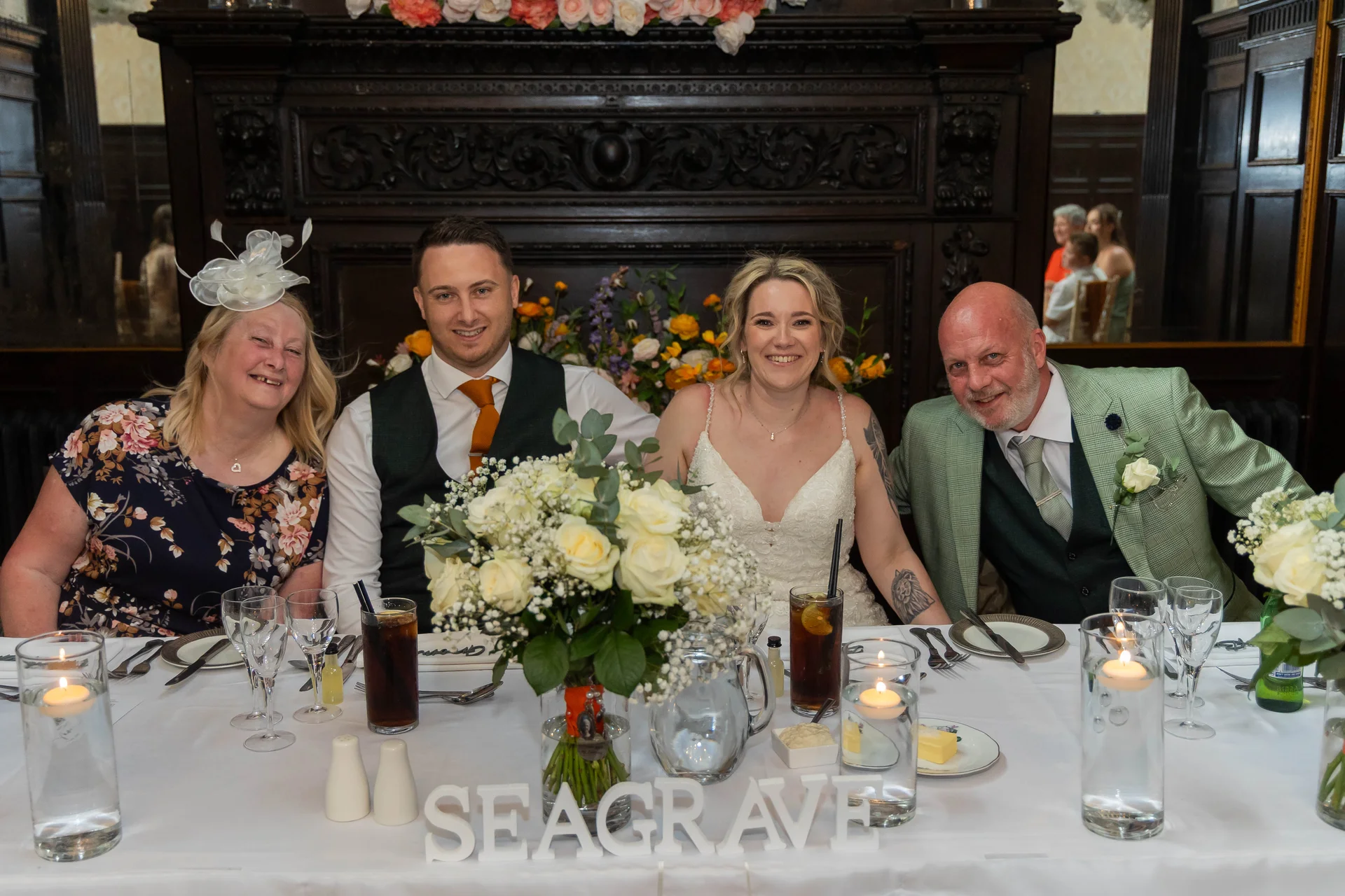 The bride and groom smile alongside parents at the top table during their wedding breakfast, featuring a centrepiece of white roses and tall candles.