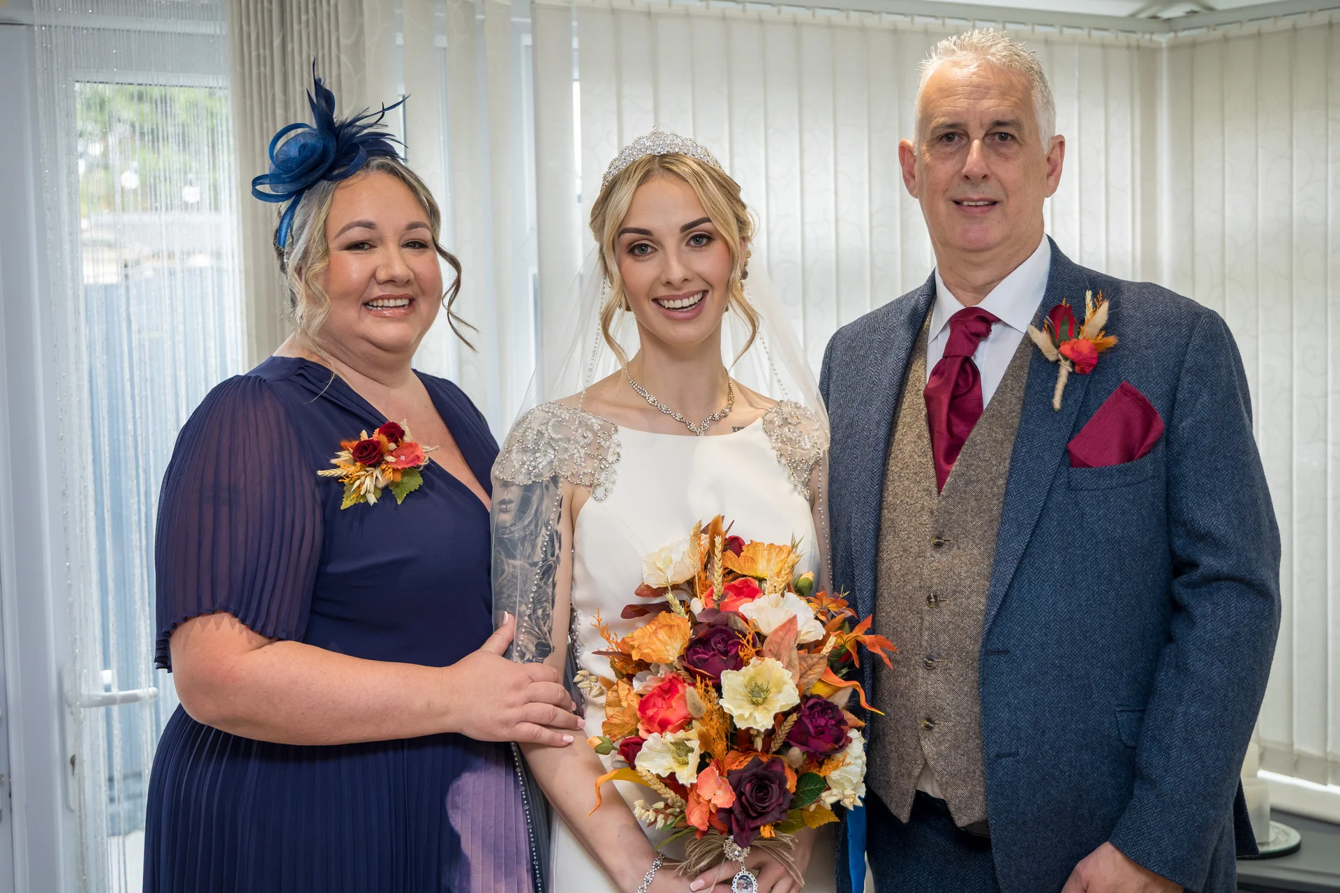 A smiling bride stands between her mother, wearing a blue dress and fascinator, and her father, wearing a tweed suit with a burnt orange buttonhole.