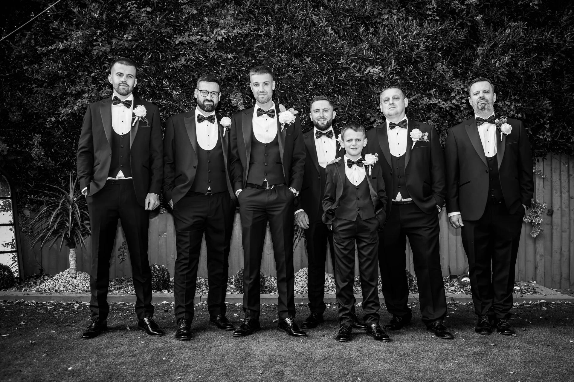 A stylish black and white portrait of the groom and his groomsmen in matching tuxedos posing outdoors in a garden.
