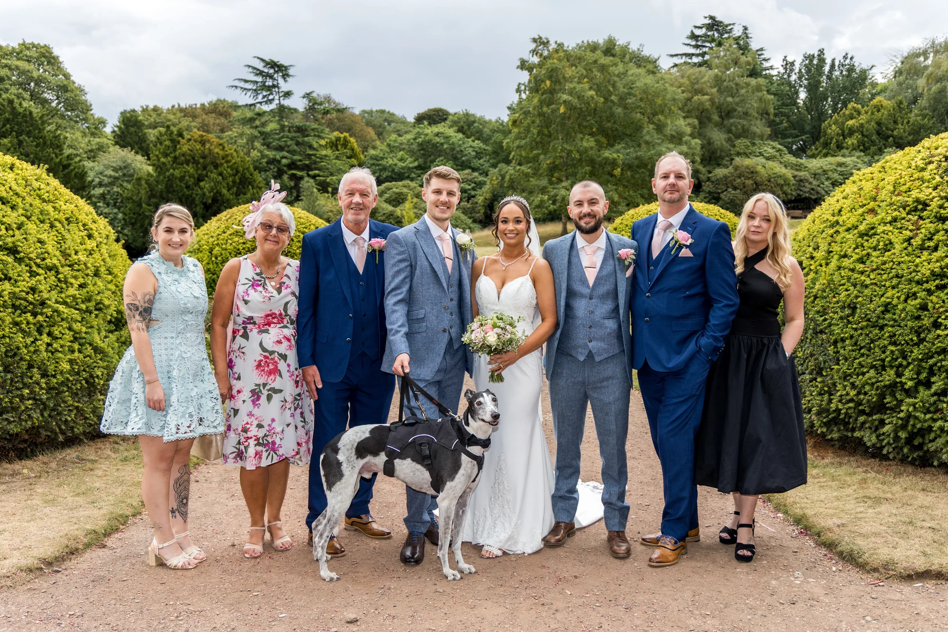 A bridal party and family members pose for a formal group portrait in a lush garden setting, accompanied by a spotted dog on a lead.