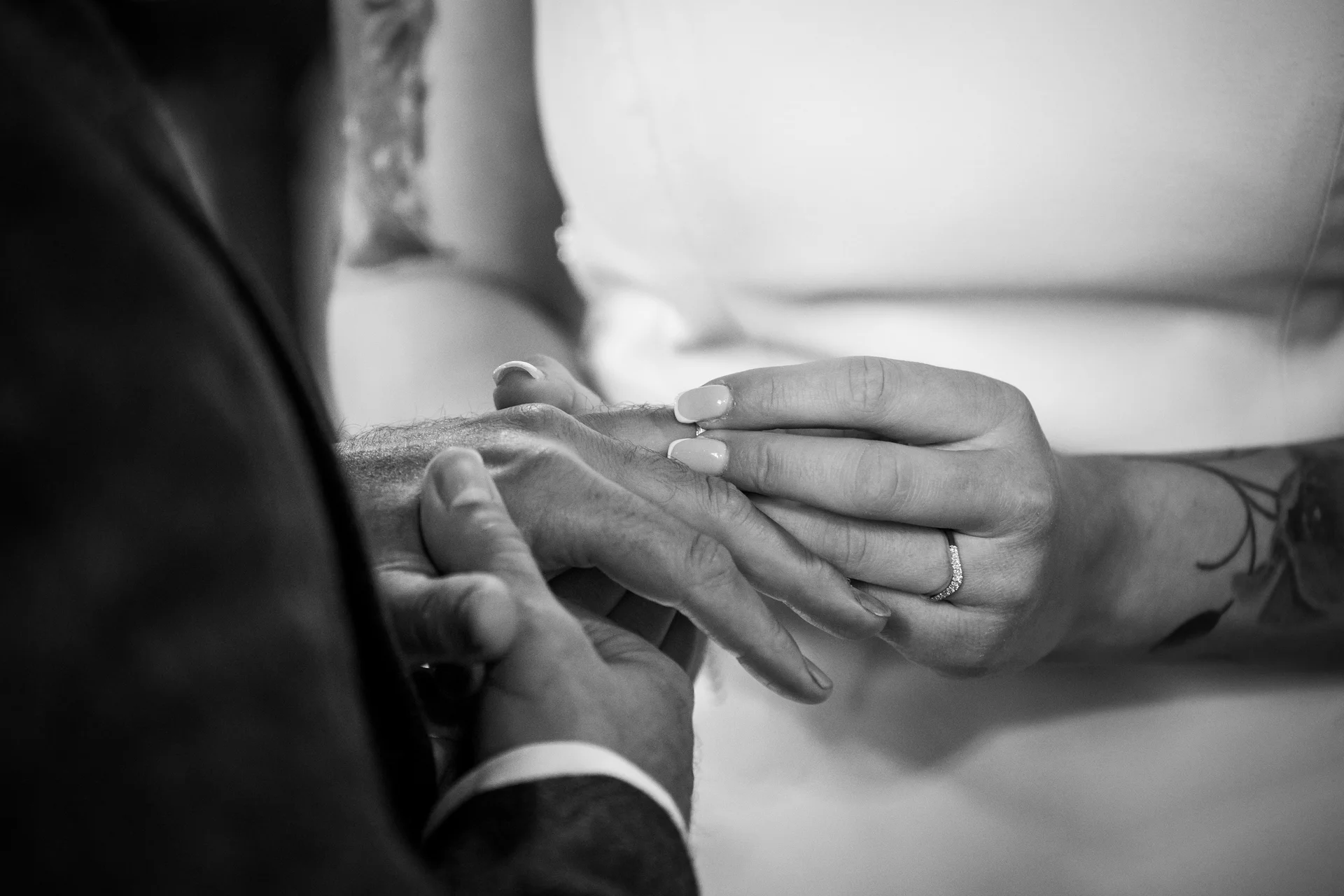 A close-up black and white shot captures the intimate moment of the bride placing the wedding ring on the groom's finger during the ceremony.
