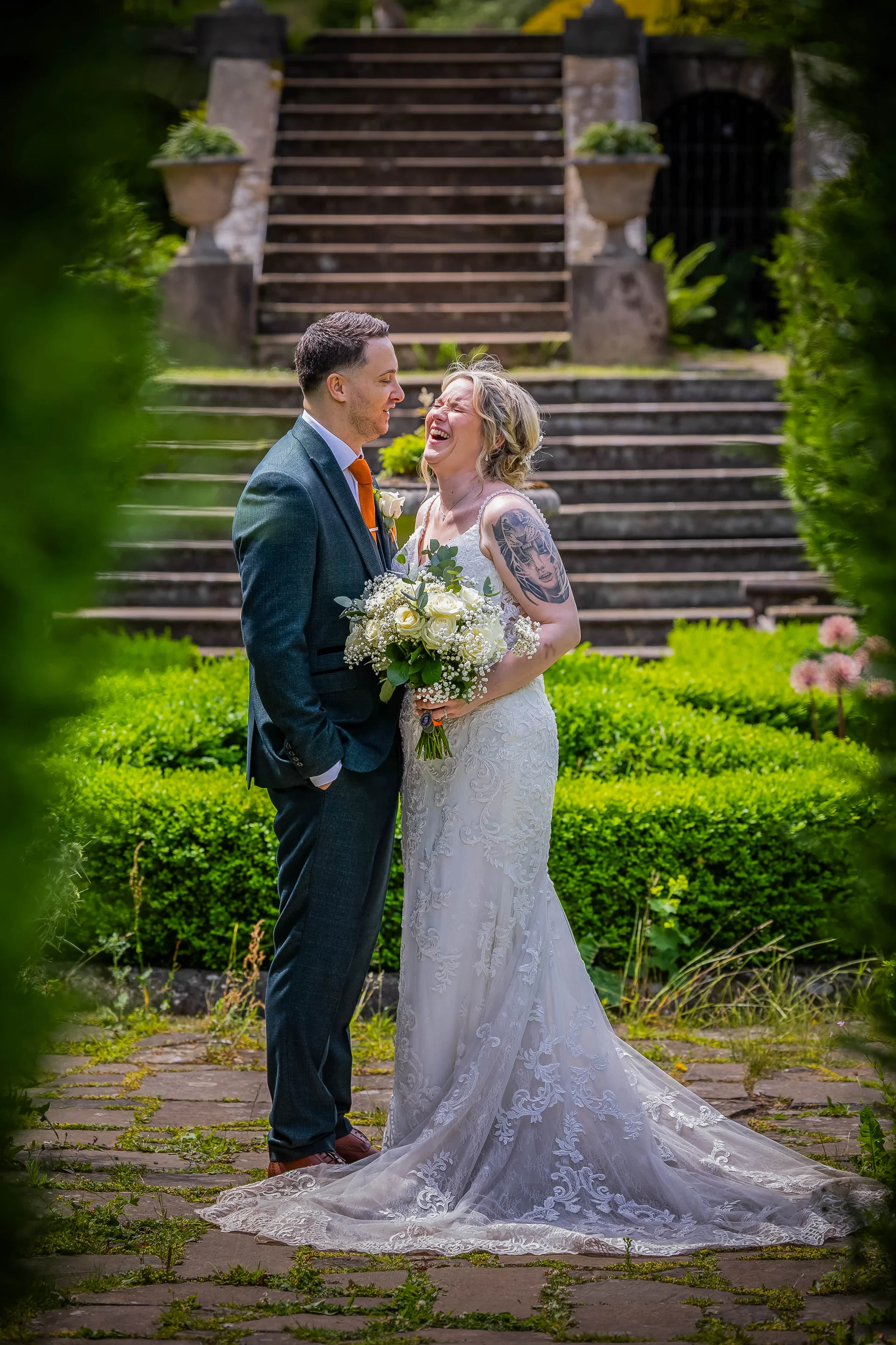 A bride with a floral tattoo on her arm laughs joyfully while holding a white rose bouquet next to her groom in a green suit outdoors.