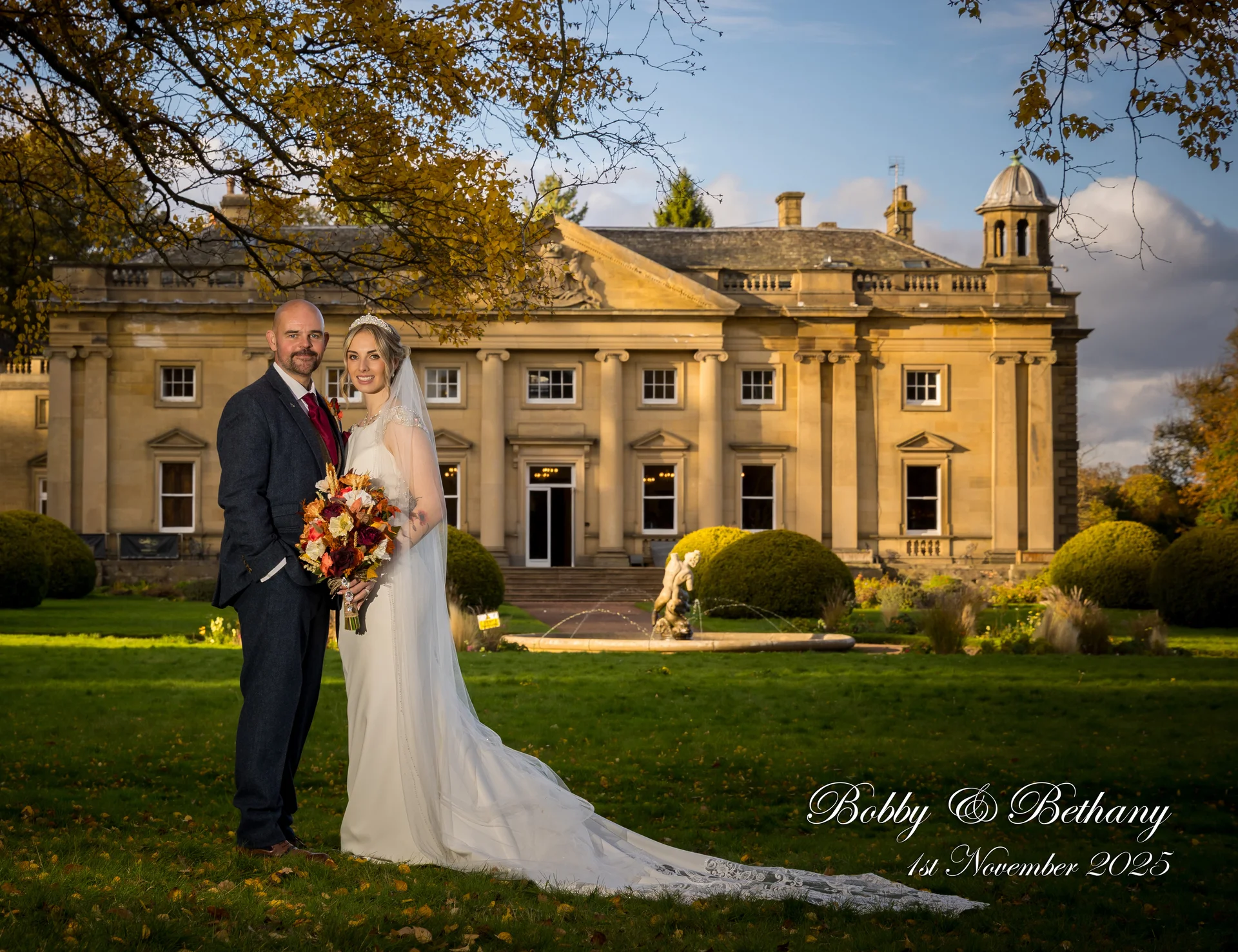 A newlywed couple poses together on a manicured lawn in front of a grand country house during autumn golden hour.