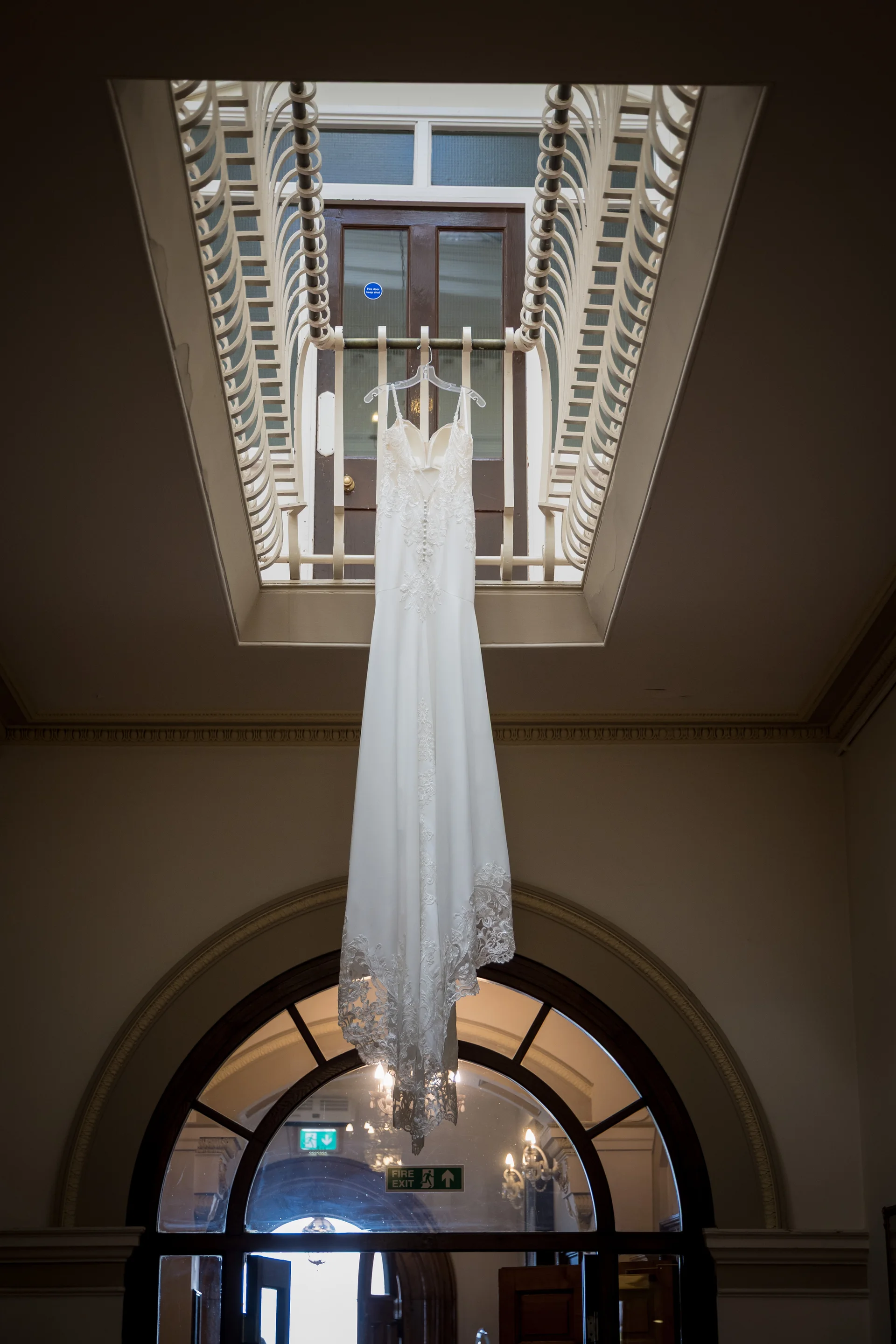 An elegant white lace wedding dress hangs vertically from an upper landing over a grand architectural staircase.