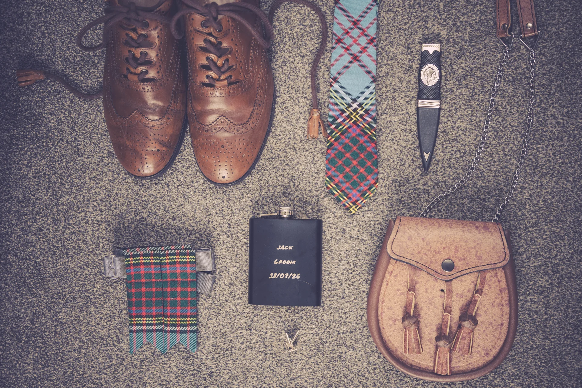 A flat lay of Scottish highland wedding attire details including brown leather brogues, a tartan tie and kilt flashes, a sgian dubh, a personalised hip flask, and a leather sporran on a grey carpet.