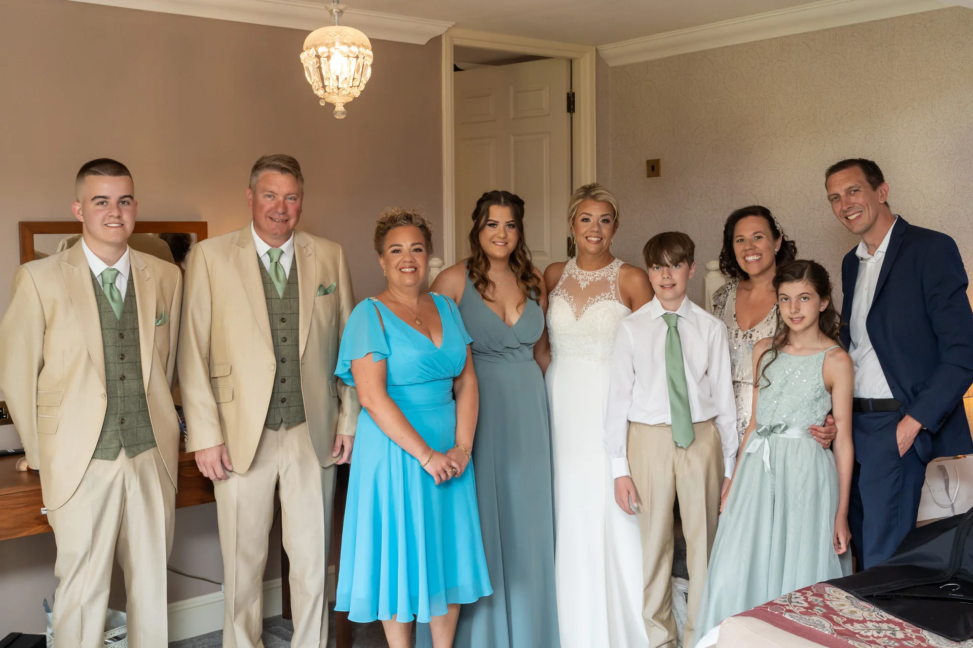A happy wedding party and family members posing together indoors, featuring the bride in a halter-neck gown alongside a groom, groomsmen in beige suits, and bridesmaids in blue and sage dresses.