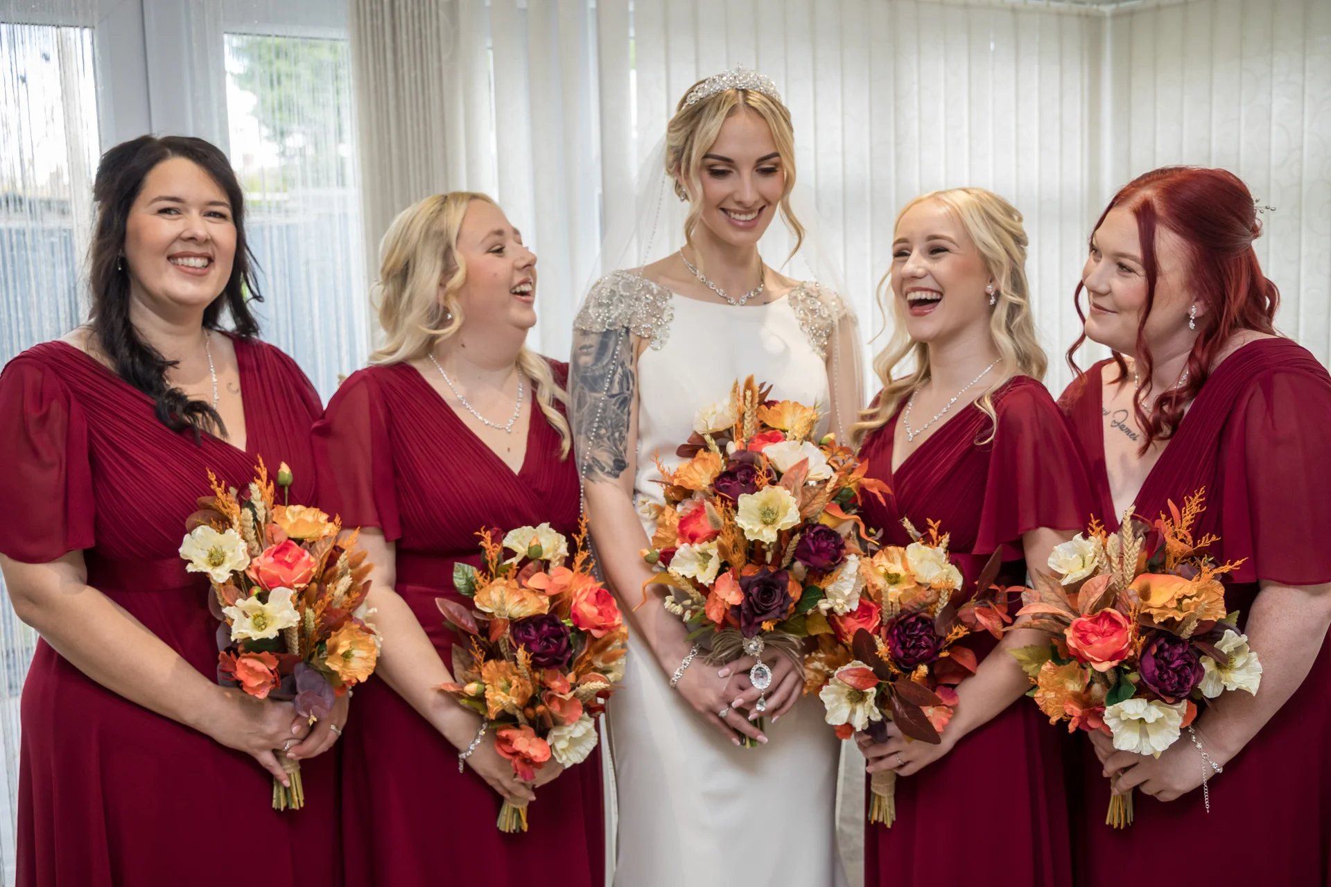 A radiant bride in a beaded white gown and tiara stands smiling among four bridesmaids wearing matching burgundy dresses and holding autumnal bouquets.