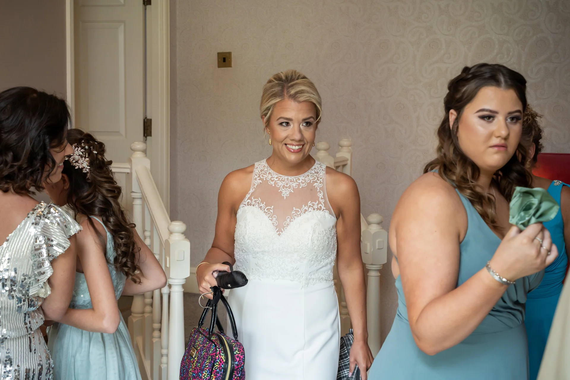 A smiling bride holding a colourful patterned handbag stands near a staircase indoors, surrounded by her bridesmaids during morning wedding preparations.
