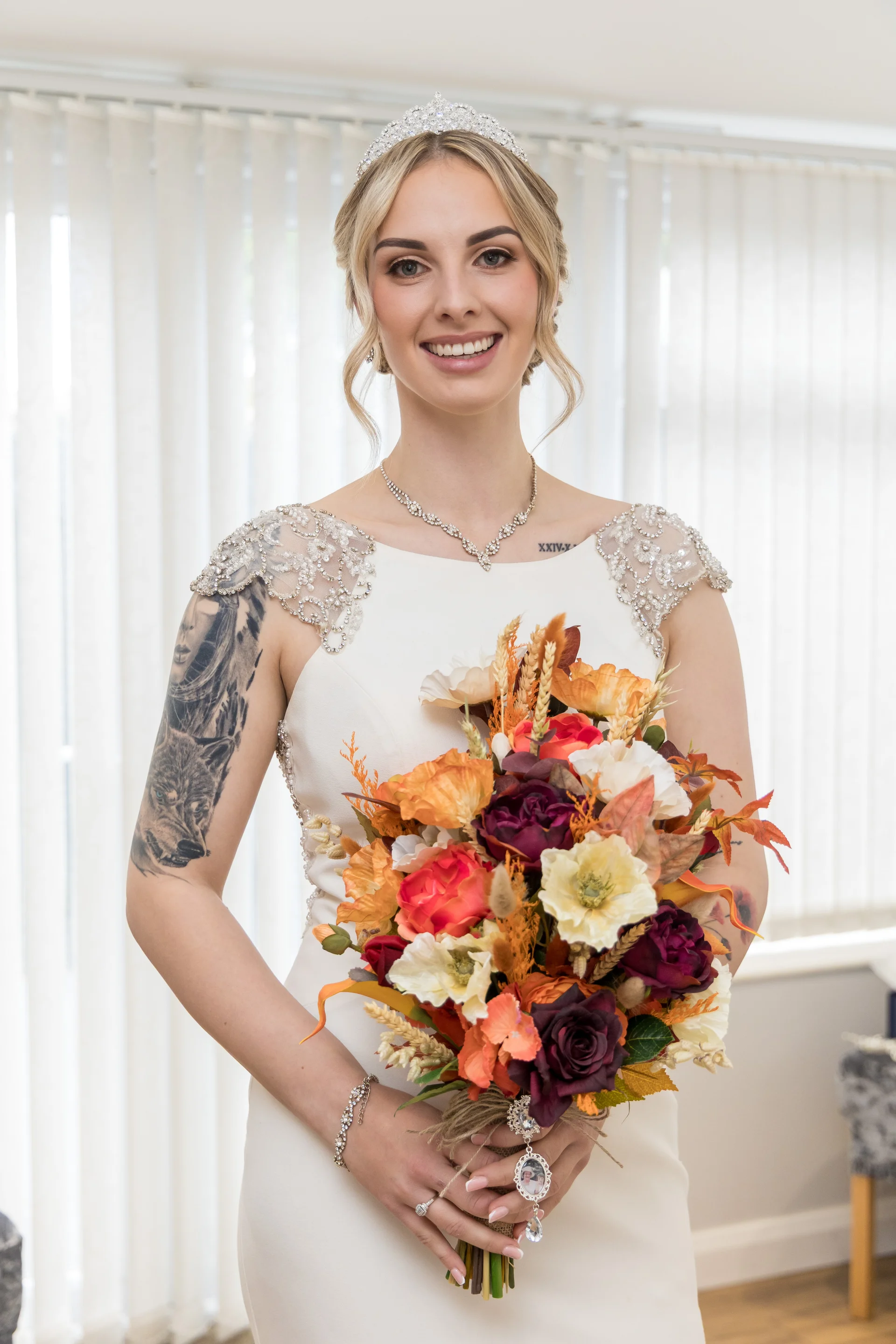 A smiling bride wearing a crystal tiara and embellished strap gown holds a vibrant autumnal bouquet featuring rich oranges and burgundy tones.