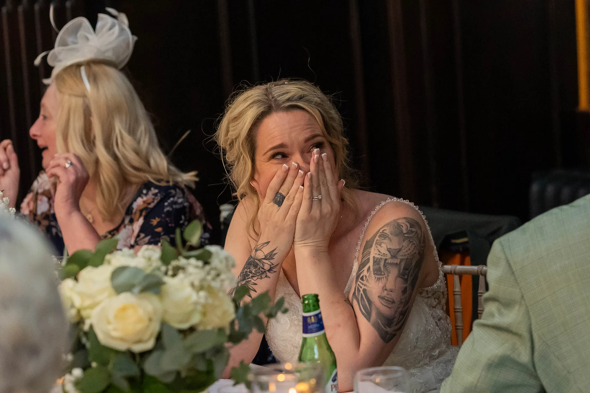 A tattooed bride sits at the reception tables with her hands covering her face, visibly emotional and crying during the wedding speeches.