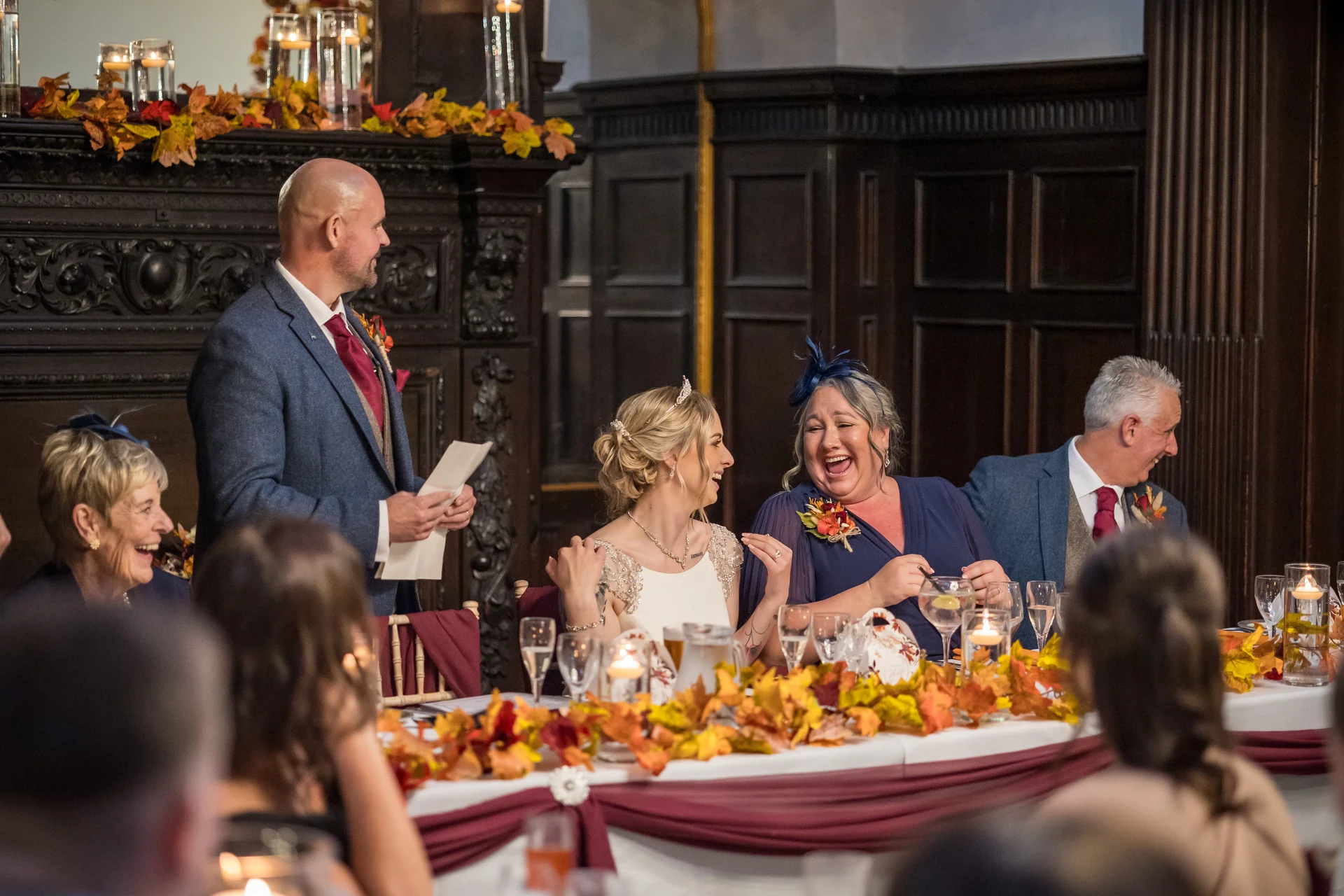 A groom delivers a speech at the top table during an autumn wedding reception, making the bride and guests laugh heartily amidst seasonal leaf decor.