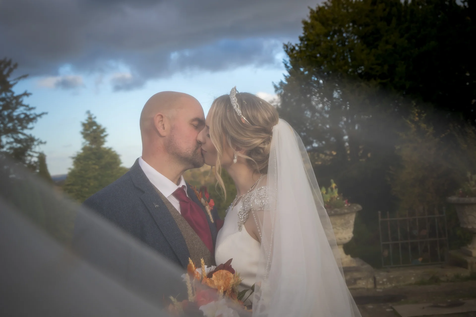 A newlywed couple shares a tender kiss outdoors under a dramatic sky, framed softly by a veil draped across the foreground.